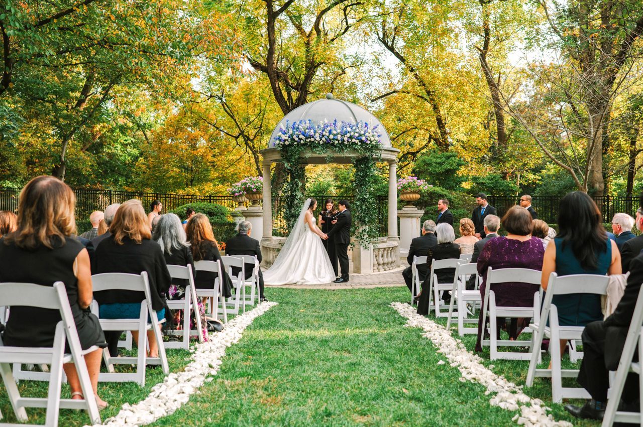 A panoramic view of an outdoor wedding ceremony setup with white chairs and floral aisle decorations in a garden setting.
