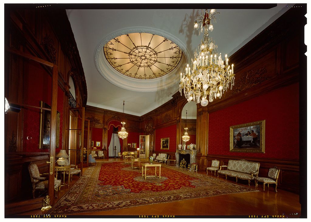 Elegant Victorian parlor with red walls, crystal chandelier, ornate ceiling medallion, and antique furnishings on Persian rug.