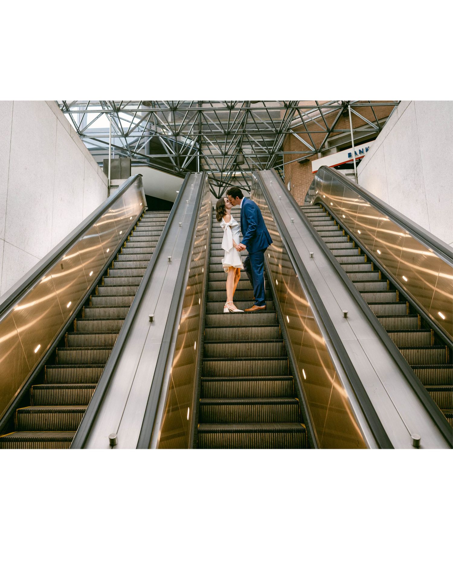Couple sharing a romantic kiss on escalators in a modern building atrium, dressed in wedding attire.