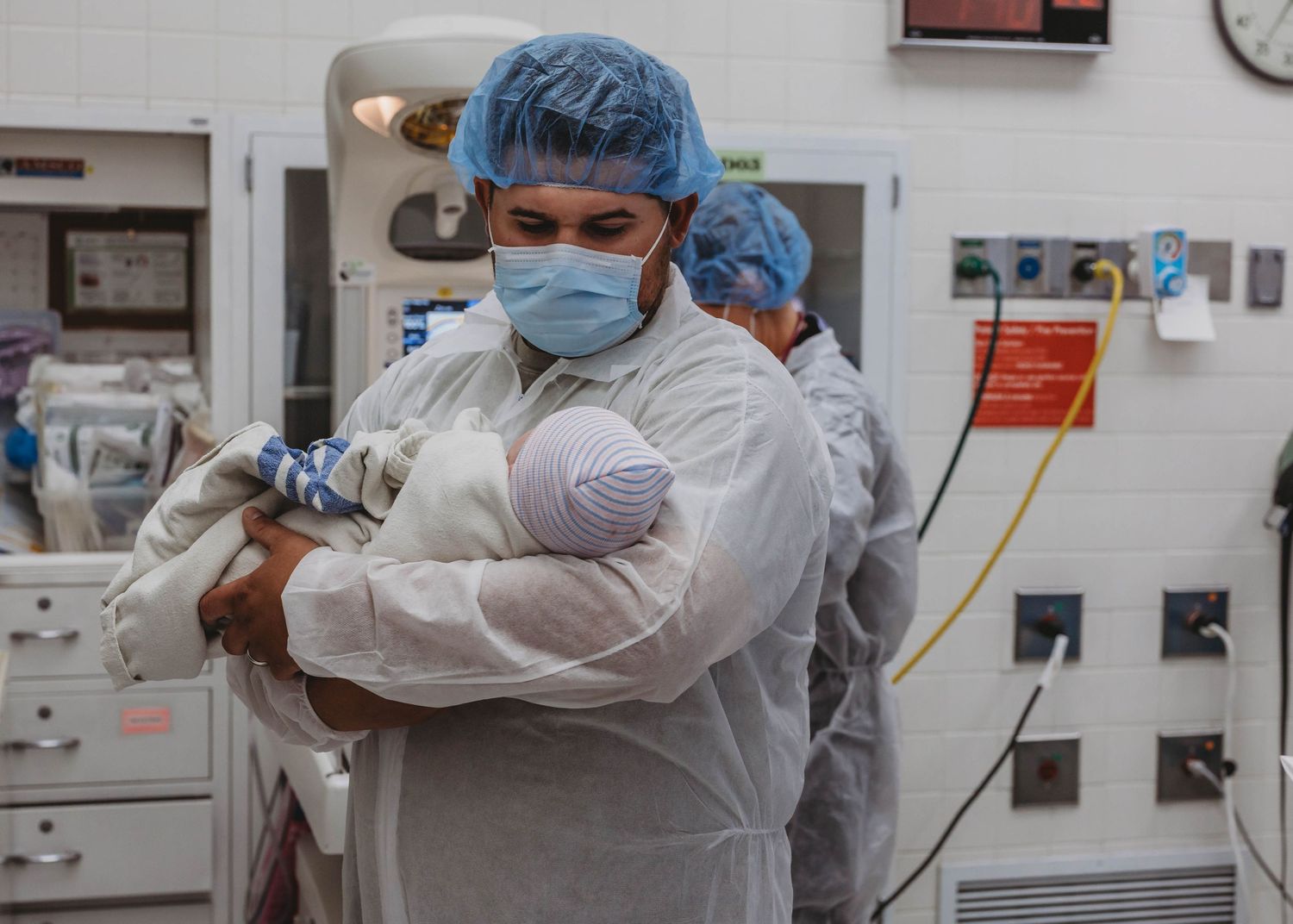 Medical professional in scrubs and mask holds a newborn baby in a hospital operating room setting.