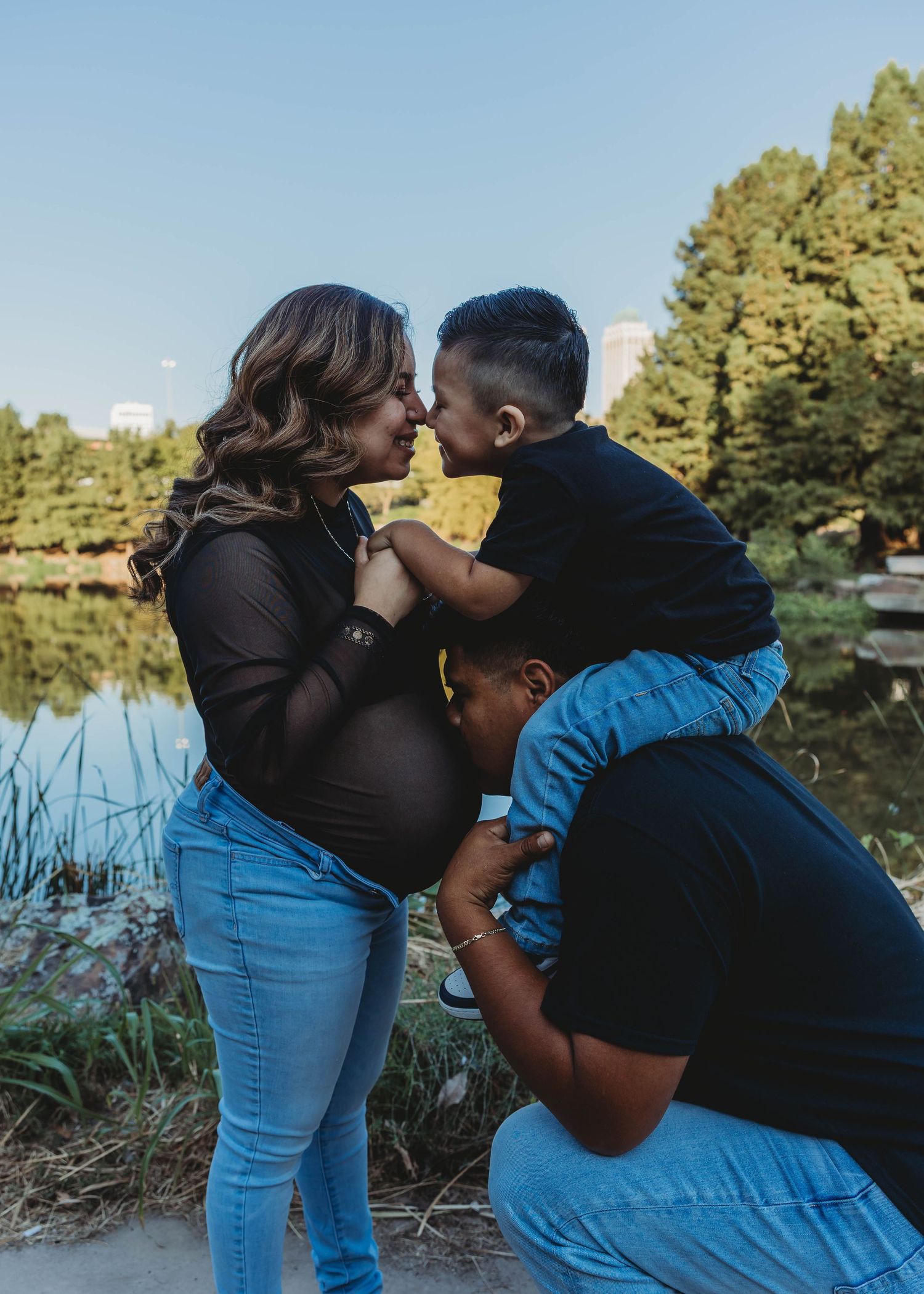 Pregnant woman shares a tender moment with her young son and partner by a serene lake in a park.