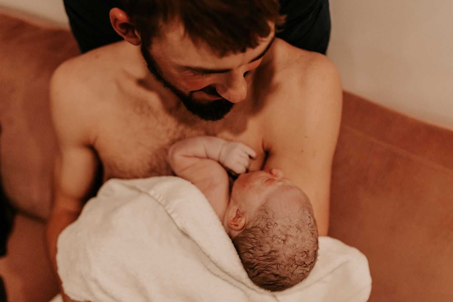 Shirtless father holding newborn baby wrapped in white towel, sharing tender skin-to-skin bonding moment.