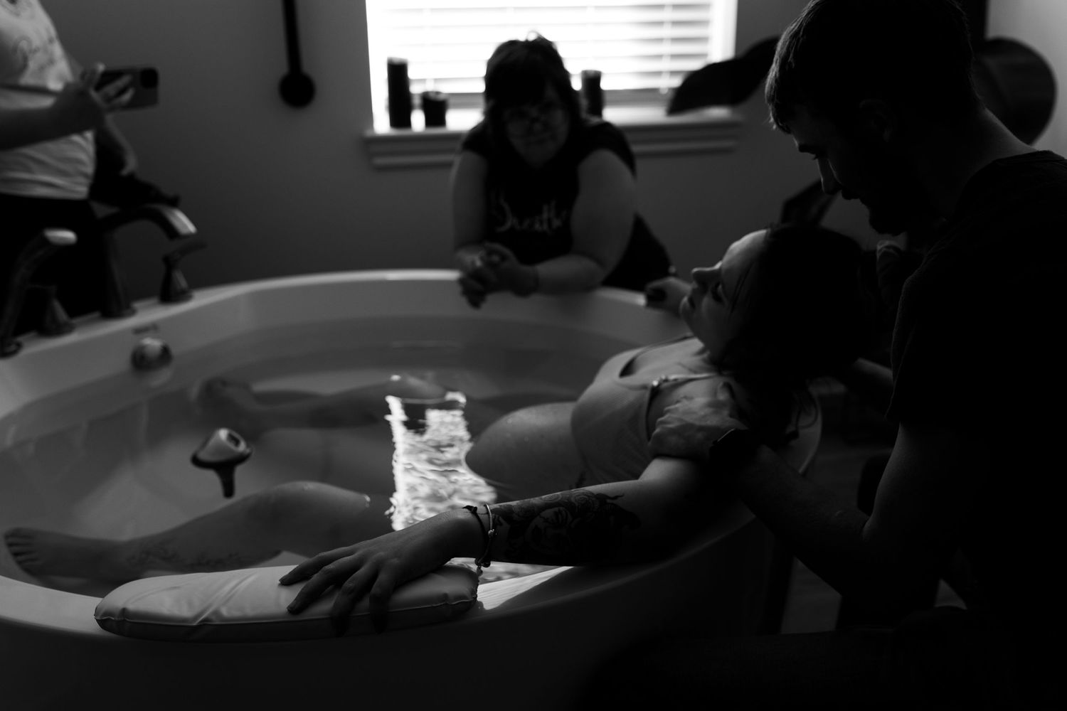 Black and white photo of a pregnant woman relaxing in a birthing tub during labor, supported by birth team at Breathe Birth and Wellness