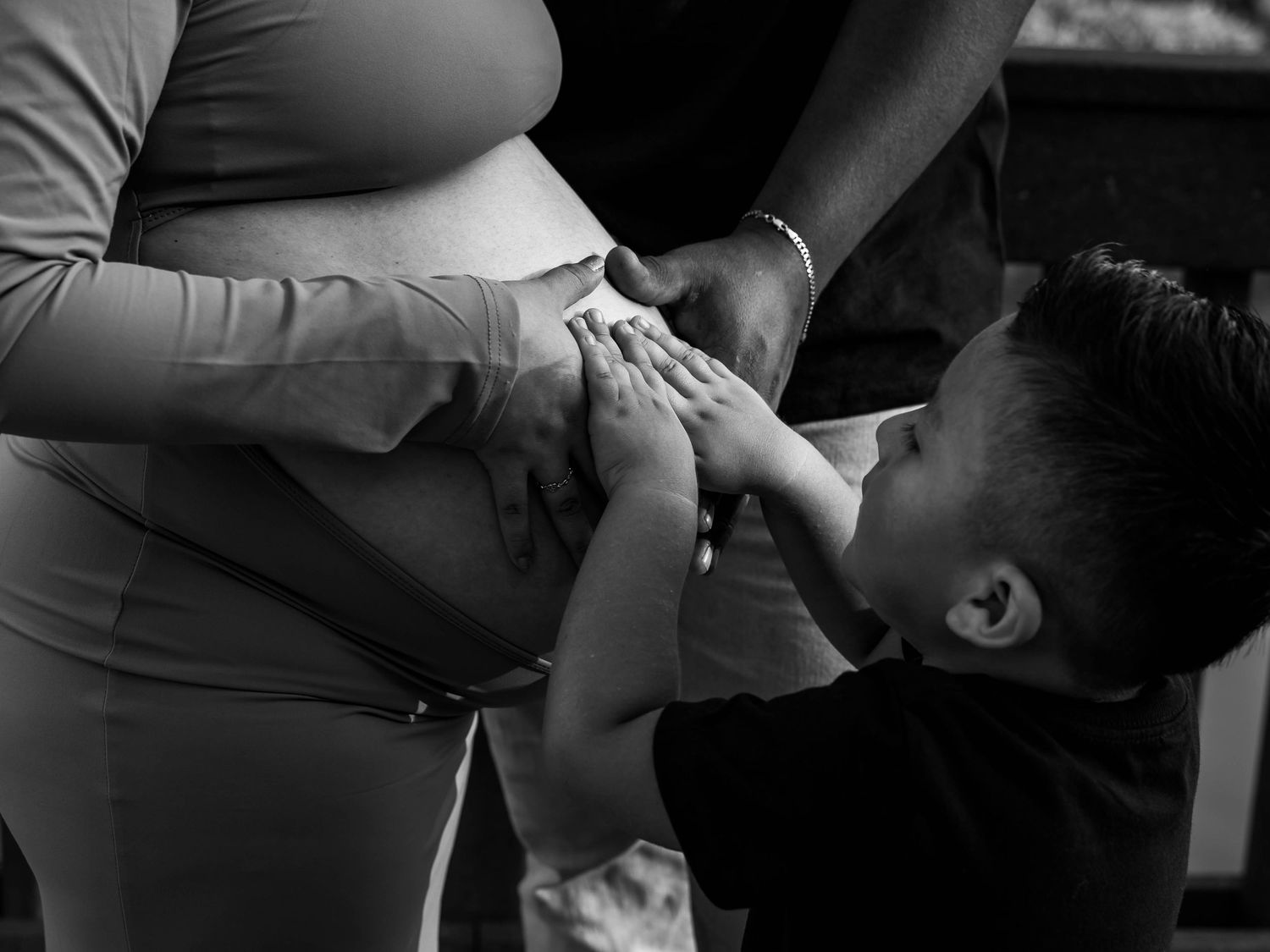 Black and white photo of a young boy kissing a pregnant woman's belly, a heartwarming family maternity moment.