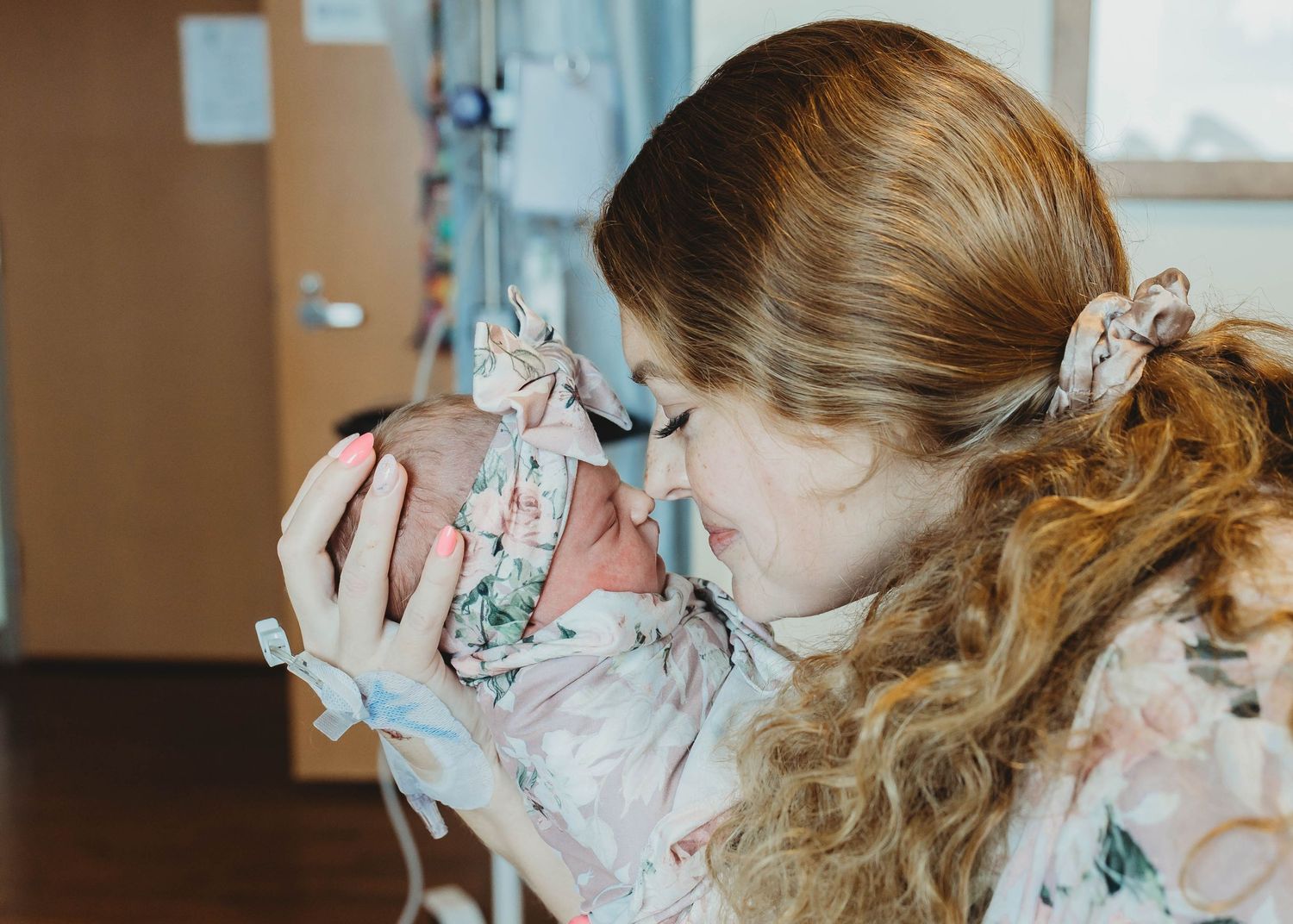 A new mother in a floral gown tenderly holds her newborn baby in a hospital room.