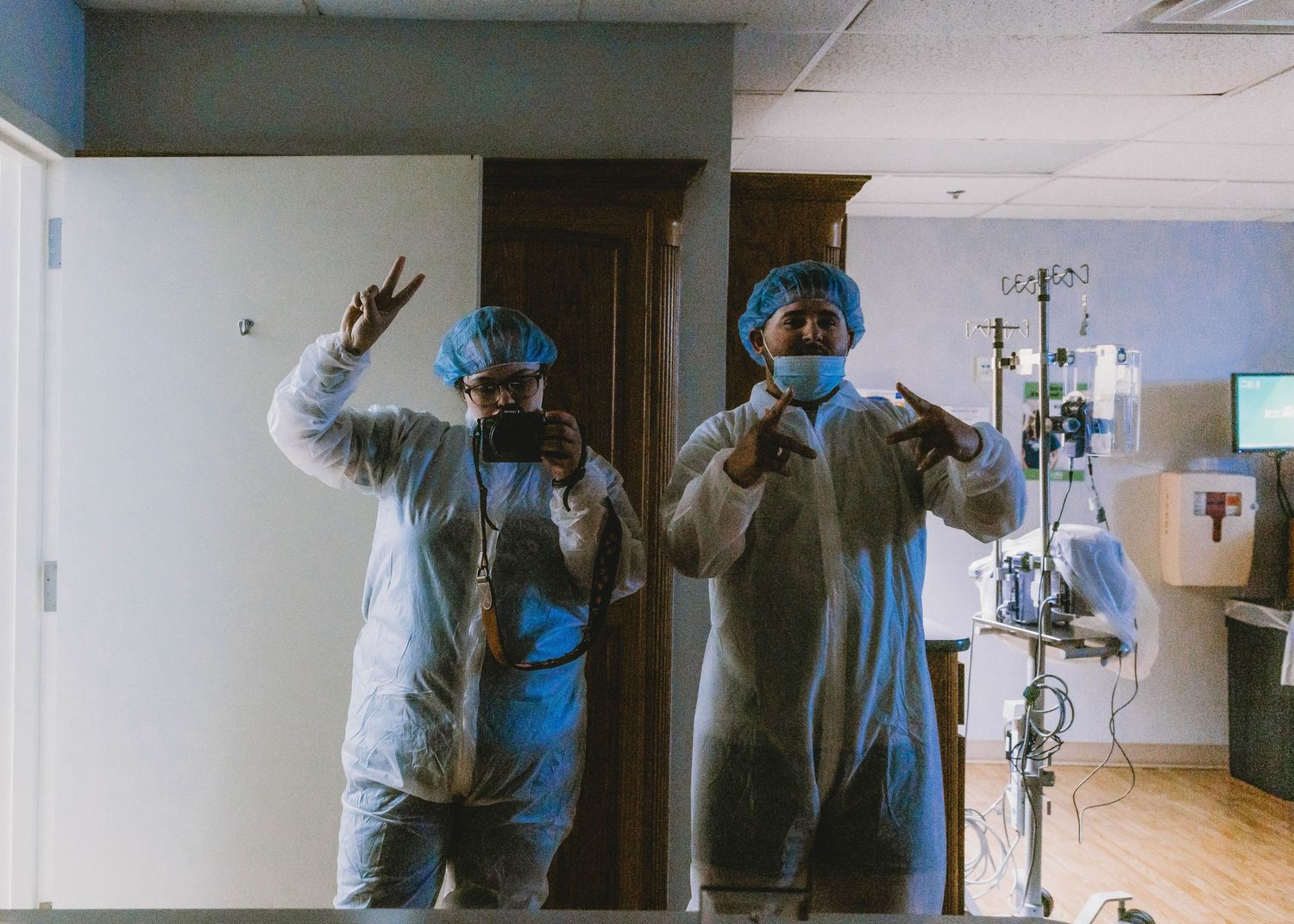 Two people in white protective gowns, blue hair caps and face masks take a mirror selfie in a hospital room with medical equipment.