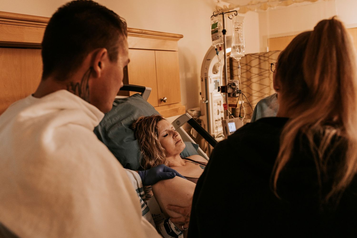 A woman in labor lies in a hospital bed surrounded by two supportive companions in a warmly lit delivery room.