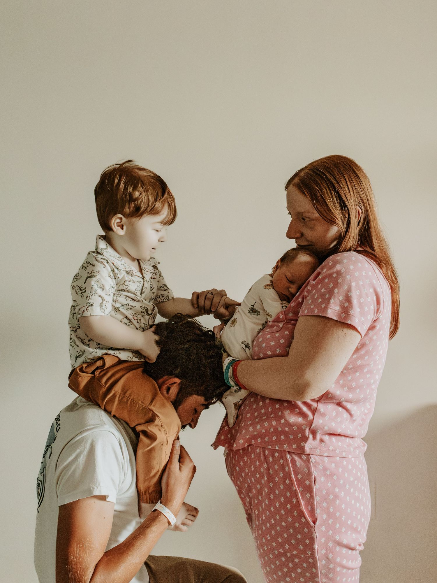 A family of four sharing a tender moment, with dad holding toddler on shoulders while mom cradles newborn baby.