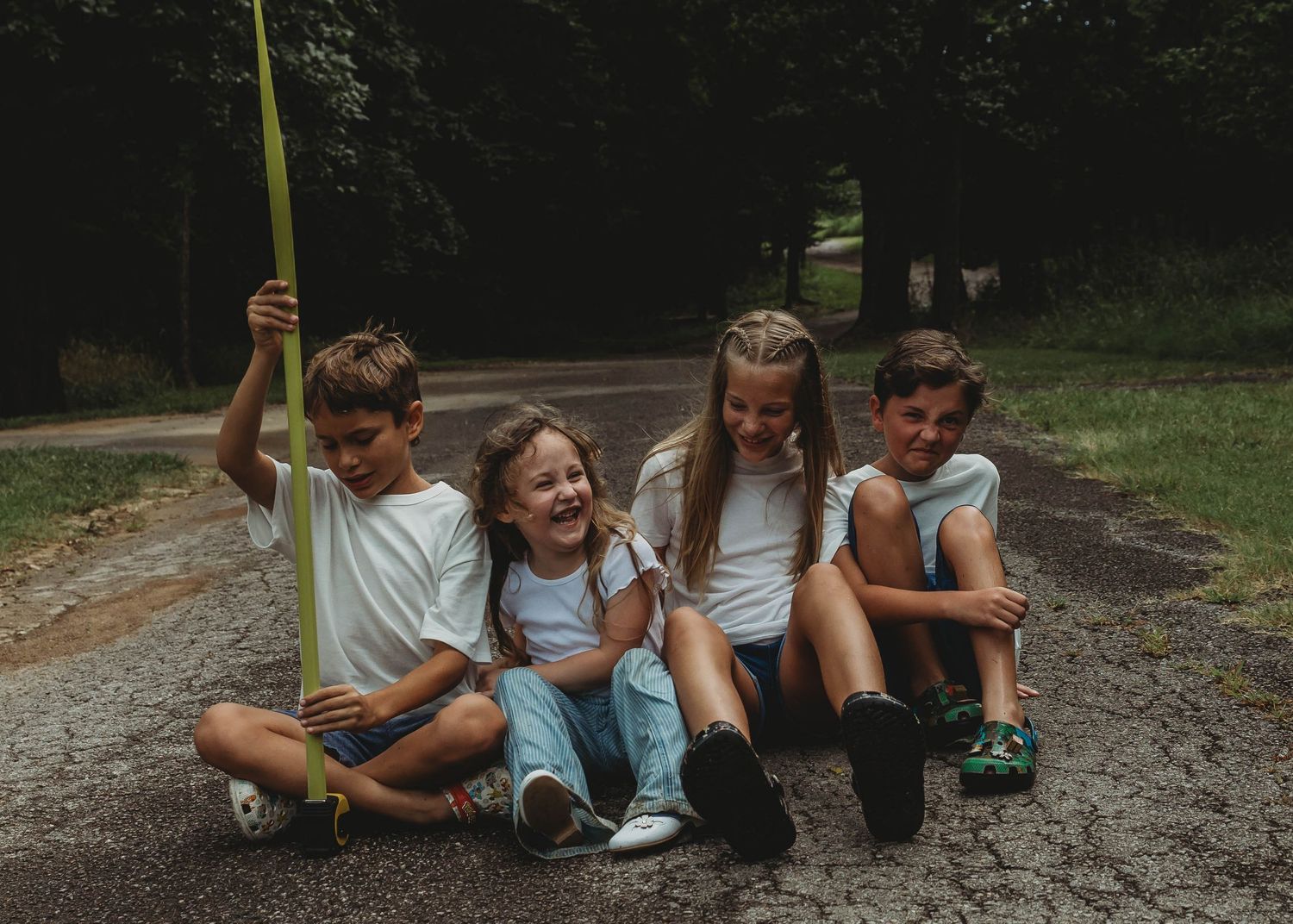 Four siblings in white shirts sitting together on a path laughing during an outdoor children photo session.