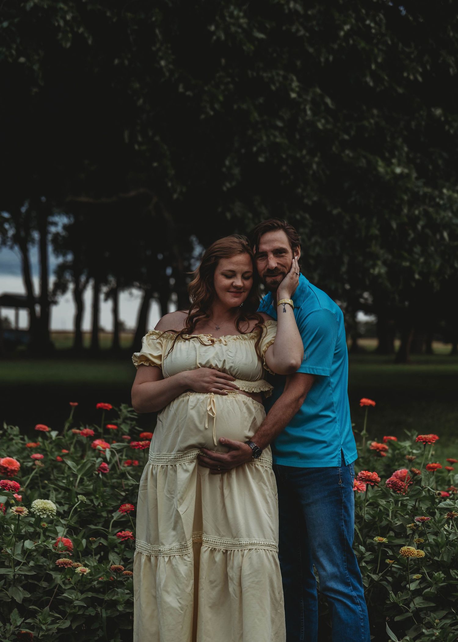 Pregnant couple posing in a flower garden, woman in cream dress cradling her bump, man in turquoise shirt embracing her.