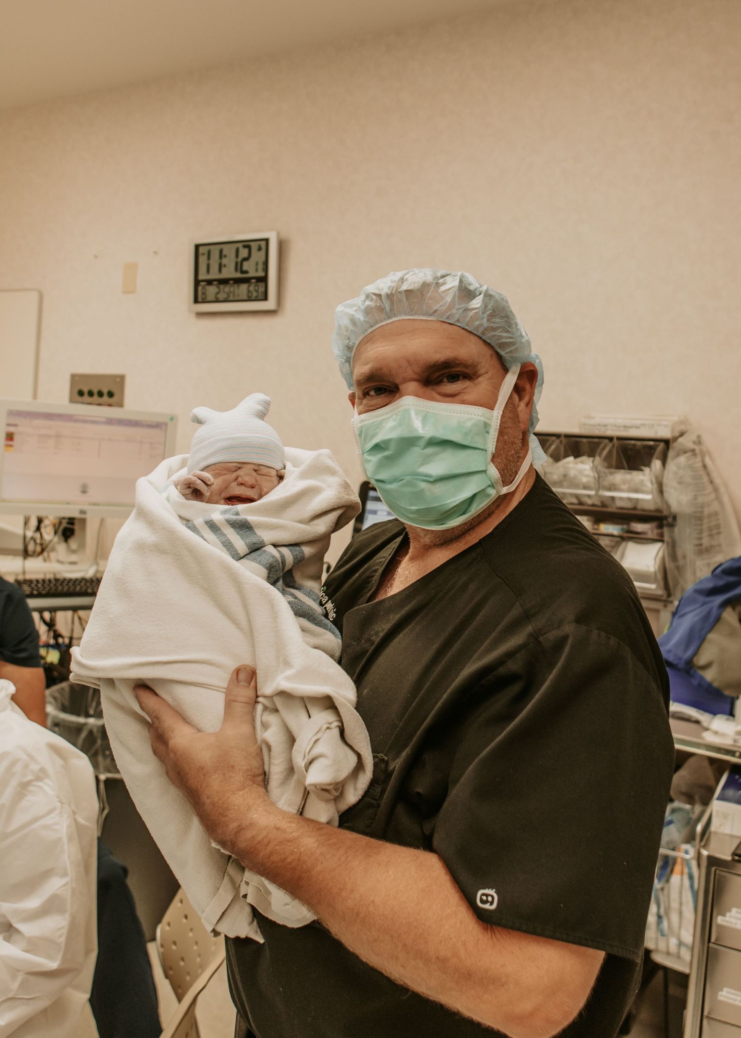 Medical professional in scrubs and surgical mask holds a swaddled newborn baby in a hospital delivery room.