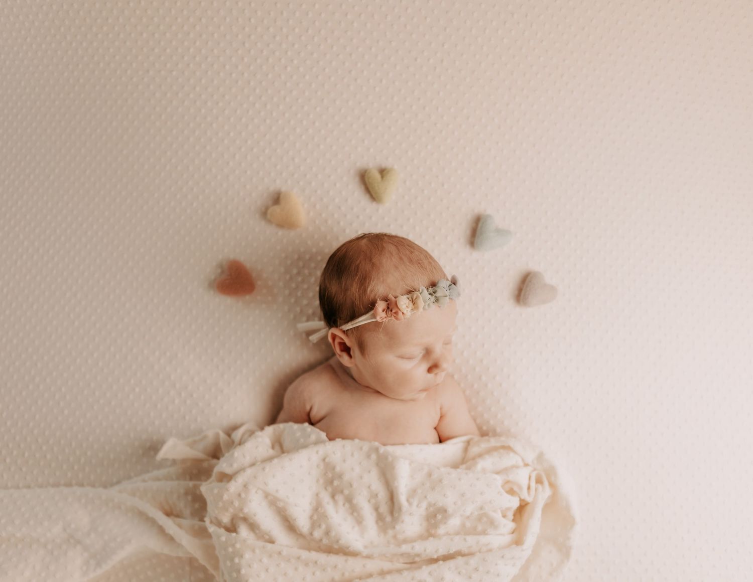 Newborn baby sleeping peacefully wrapped in cream blanket surrounded by colorful felt hearts on white background.