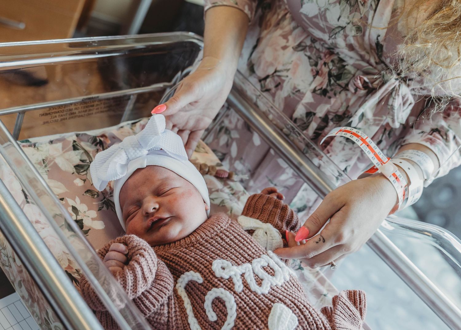 Newborn baby in knit outfit lying in hospital bassinet with mother gently touching hand.