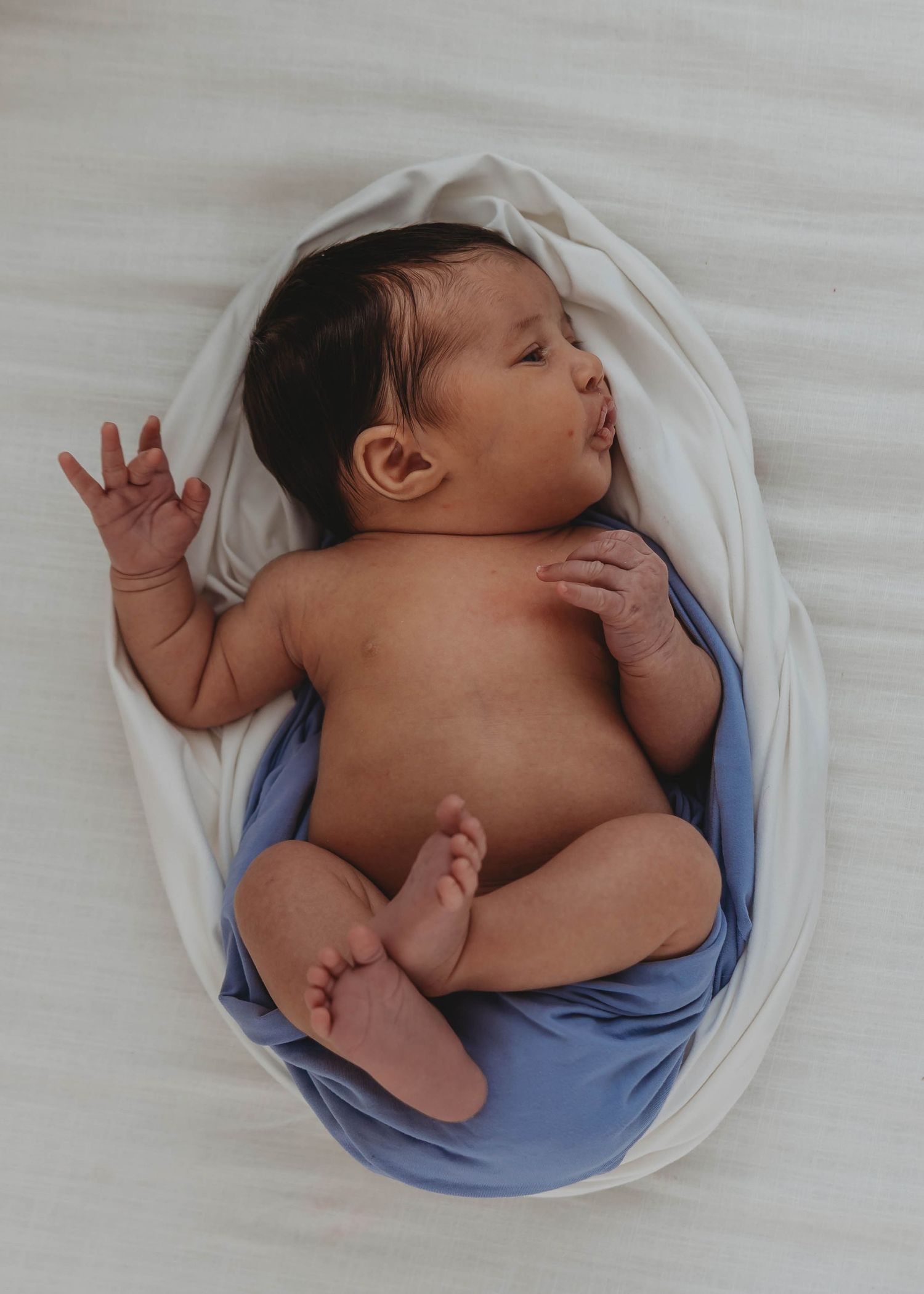Newborn baby lying in a white and blue swaddle blanket with one hand raised, eyes closed peacefully.