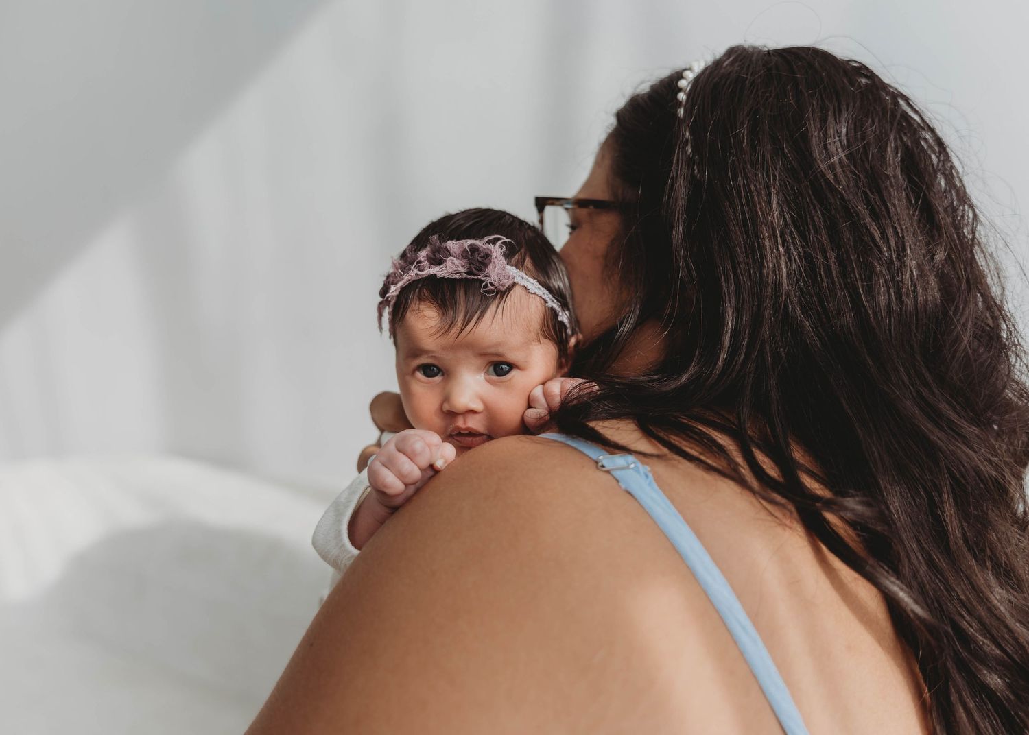 A mother tenderly holds her newborn baby wearing a pink floral headband against a soft white background in an Oklahoma studio.