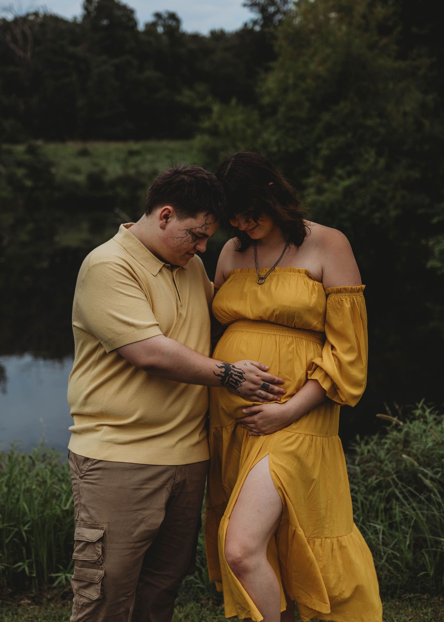 Expecting couple in yellow outfits tenderly cradling baby bump near a pond during outdoor maternity photo session.