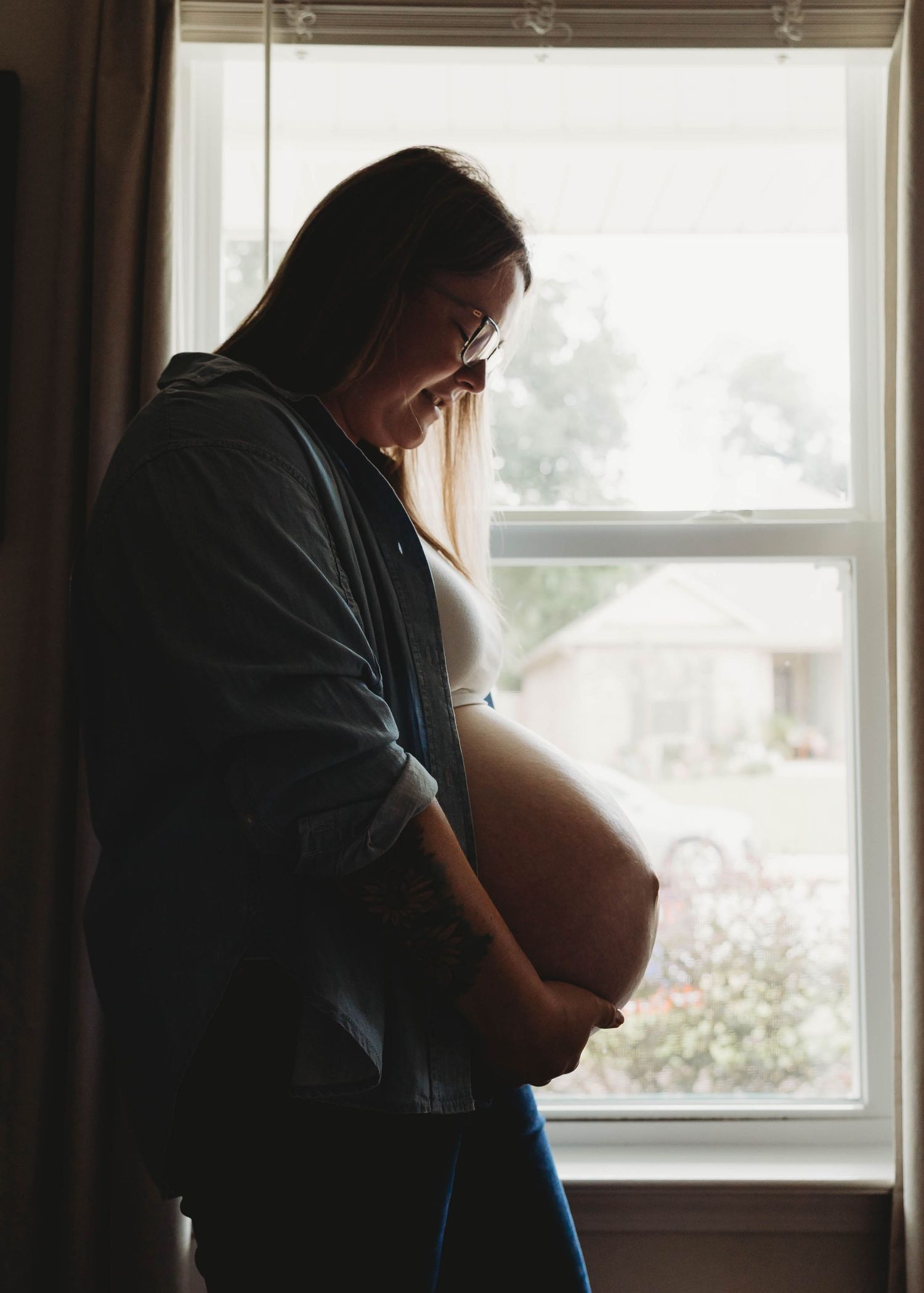 Pregnant woman standing by window cradling her bare baby bump in soft natural light during maternity photo session.