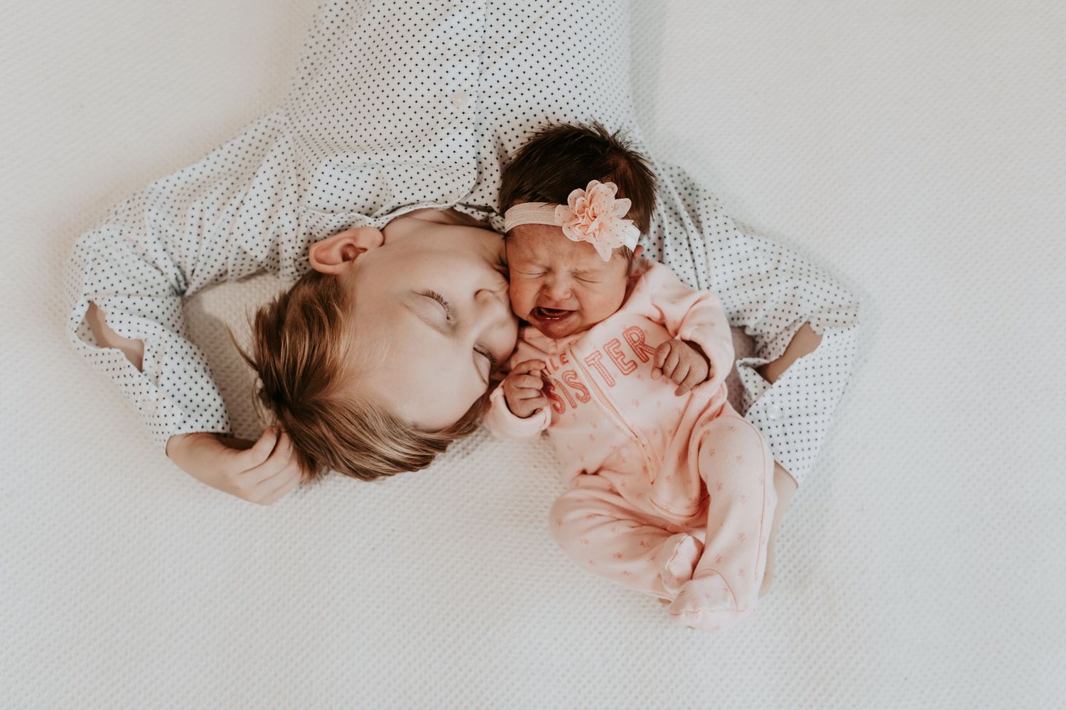 Older sibling kissing newborn baby dressed in pink on a white blanket, sweet newborn lifestyle photography.