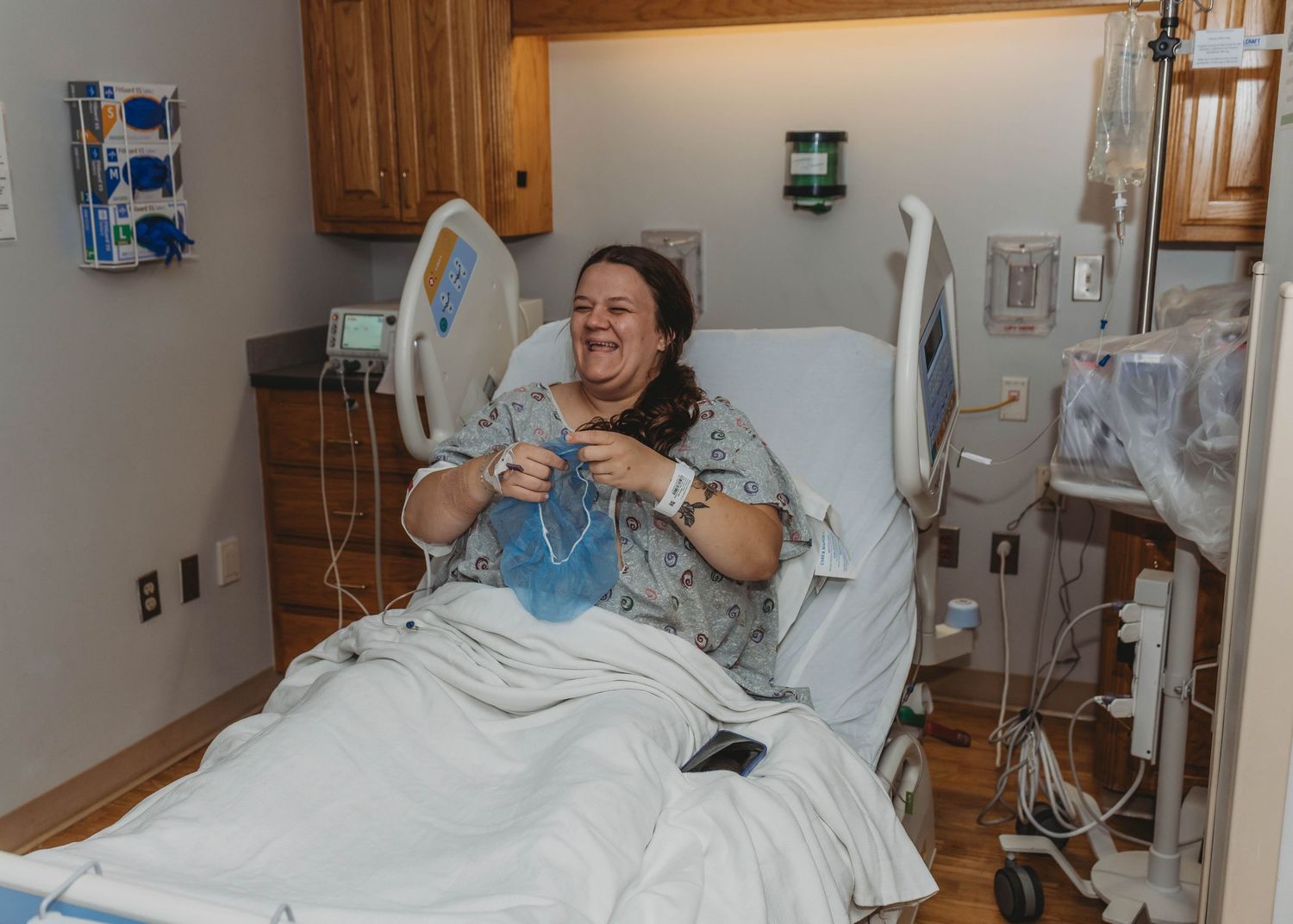 Smiling woman in a hospital gown sits up in a birthing room bed, holding a blue hair cap preparing for her c-section in Claremore, OK