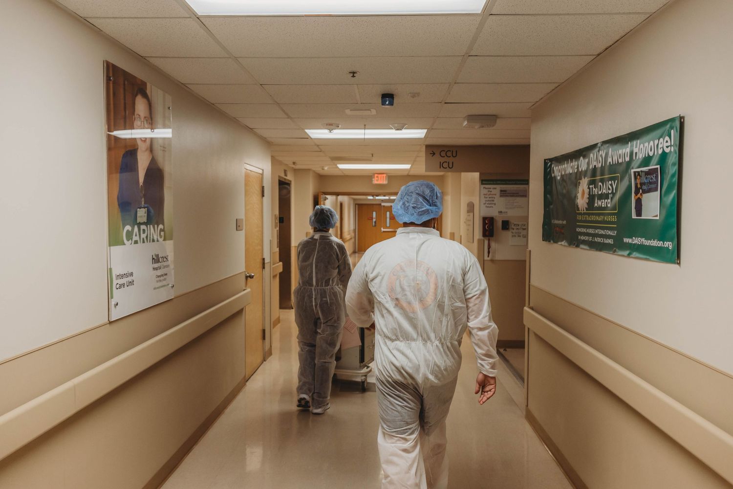 Two healthcare workers in protective gowns and surgical caps walk down a hospital corridor lined with wall posters and handrails.