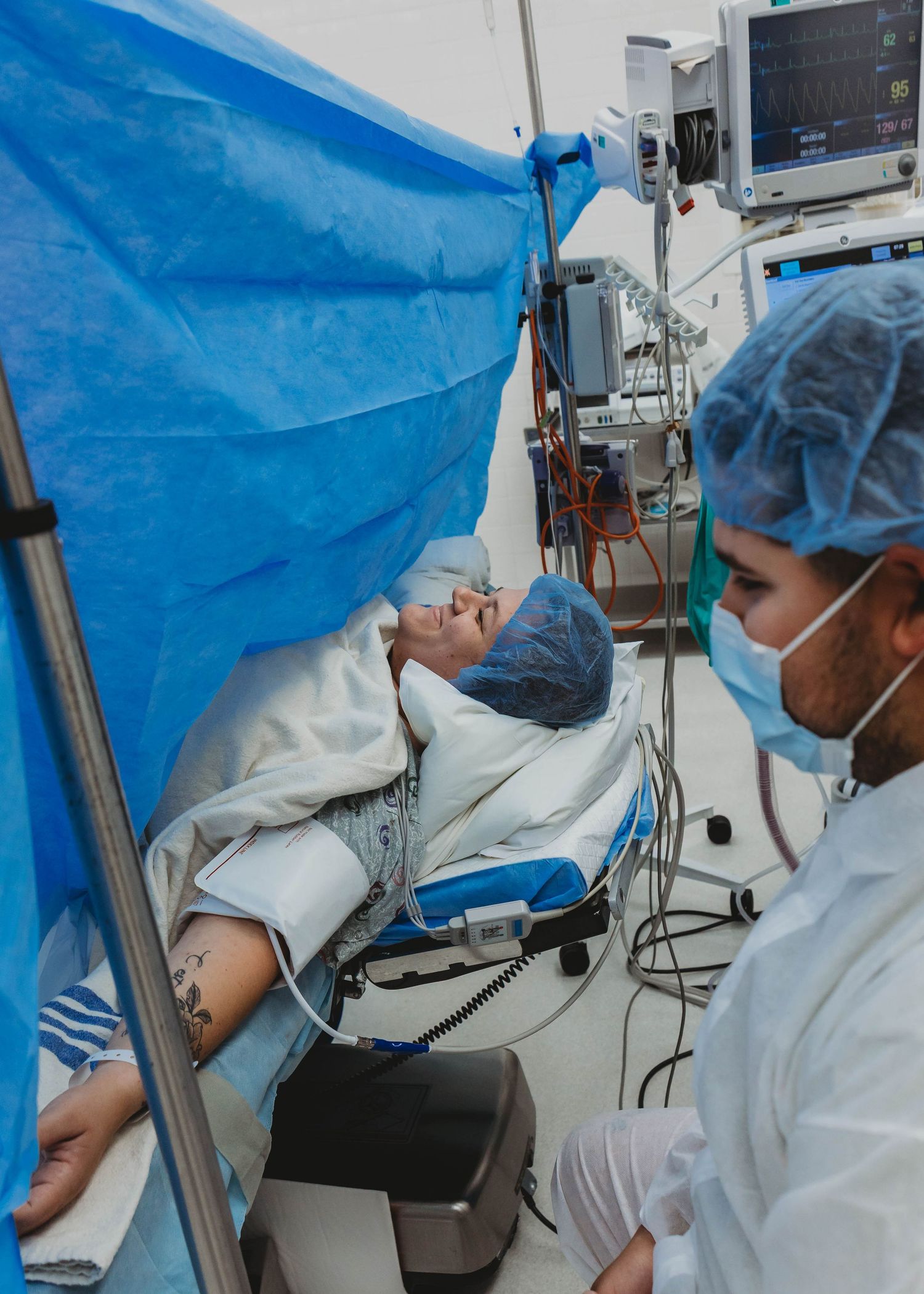 Patient in a surgical cap lies on an operating table behind a blue drape while a masked medical worker in scrubs stands beside monitors.