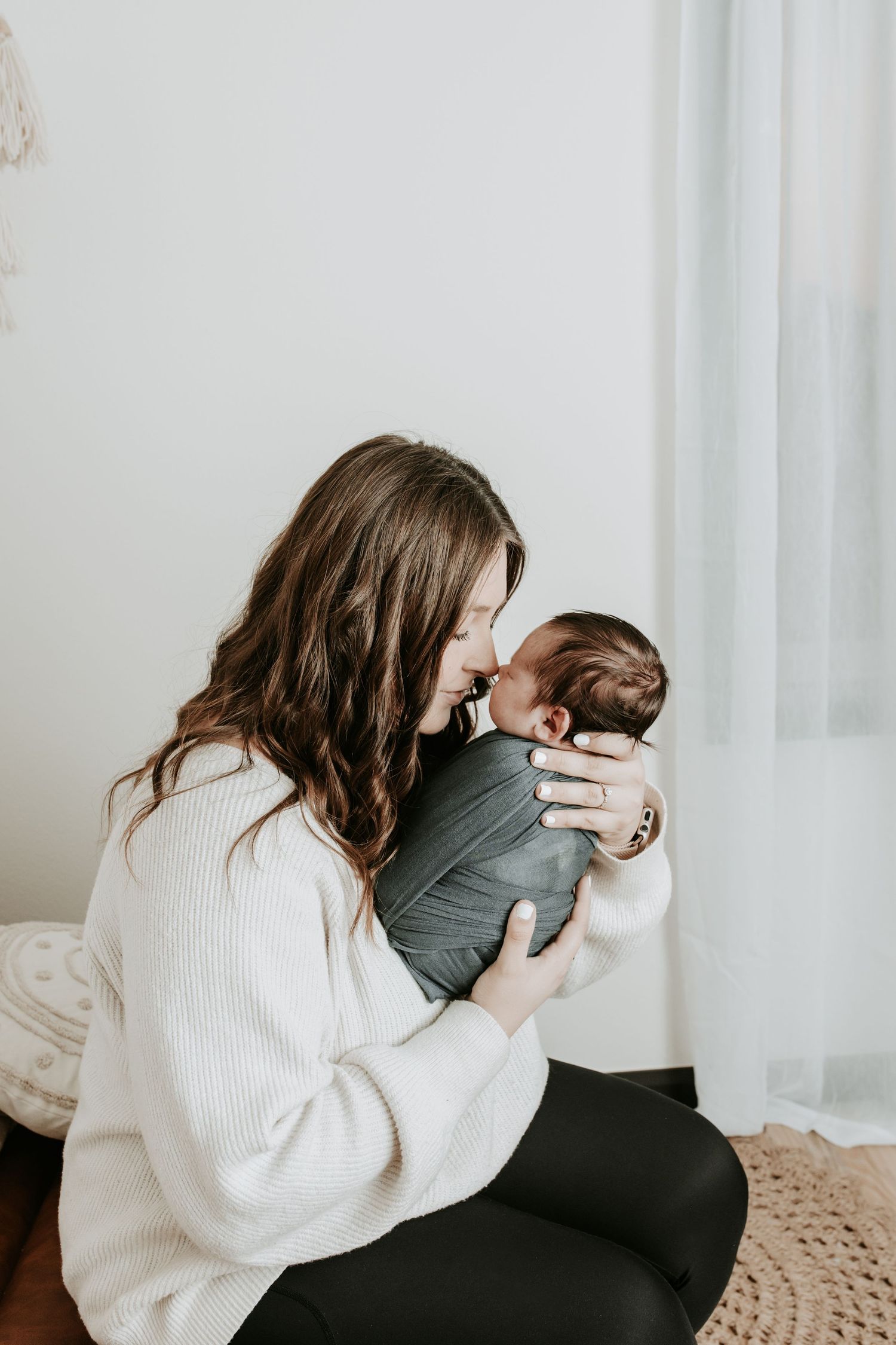A young mother in a white sweater tenderly holds her swaddled newborn baby close to her face near a bright window.