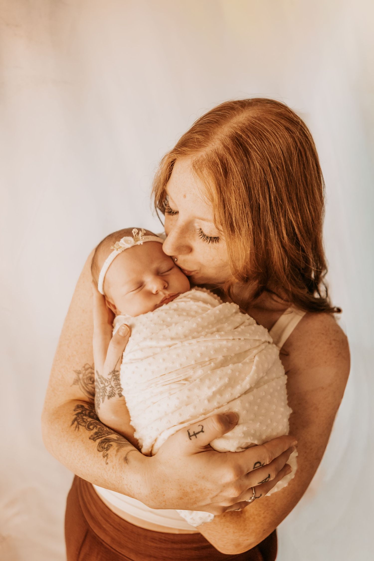 Red-haired mother with tattoos kissing her swaddled newborn baby wrapped in cream blanket.