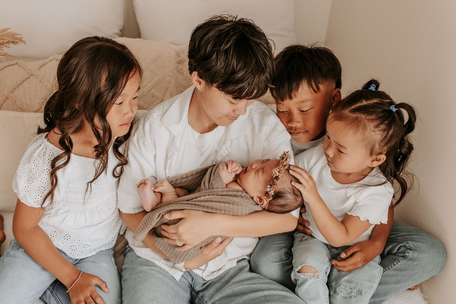 Four siblings in white shirts cuddle together on a bed, lovingly holding a newborn baby wrapped in a pink blanket in Coweta Oklahoma