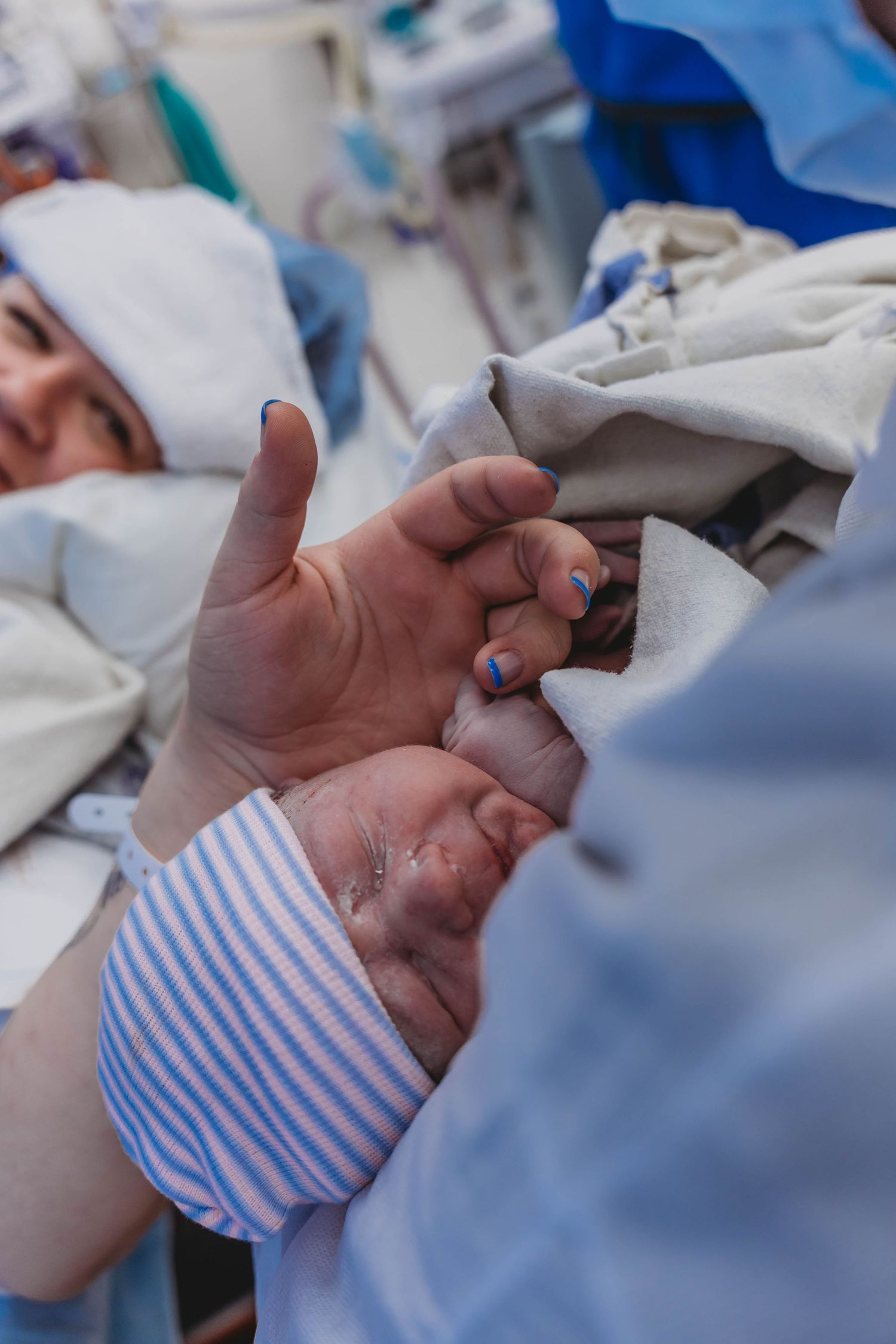 Newborn baby in a striped hospital beanie held against a blue gown while a parent's hand gives a thumbs up from a hospital bed.