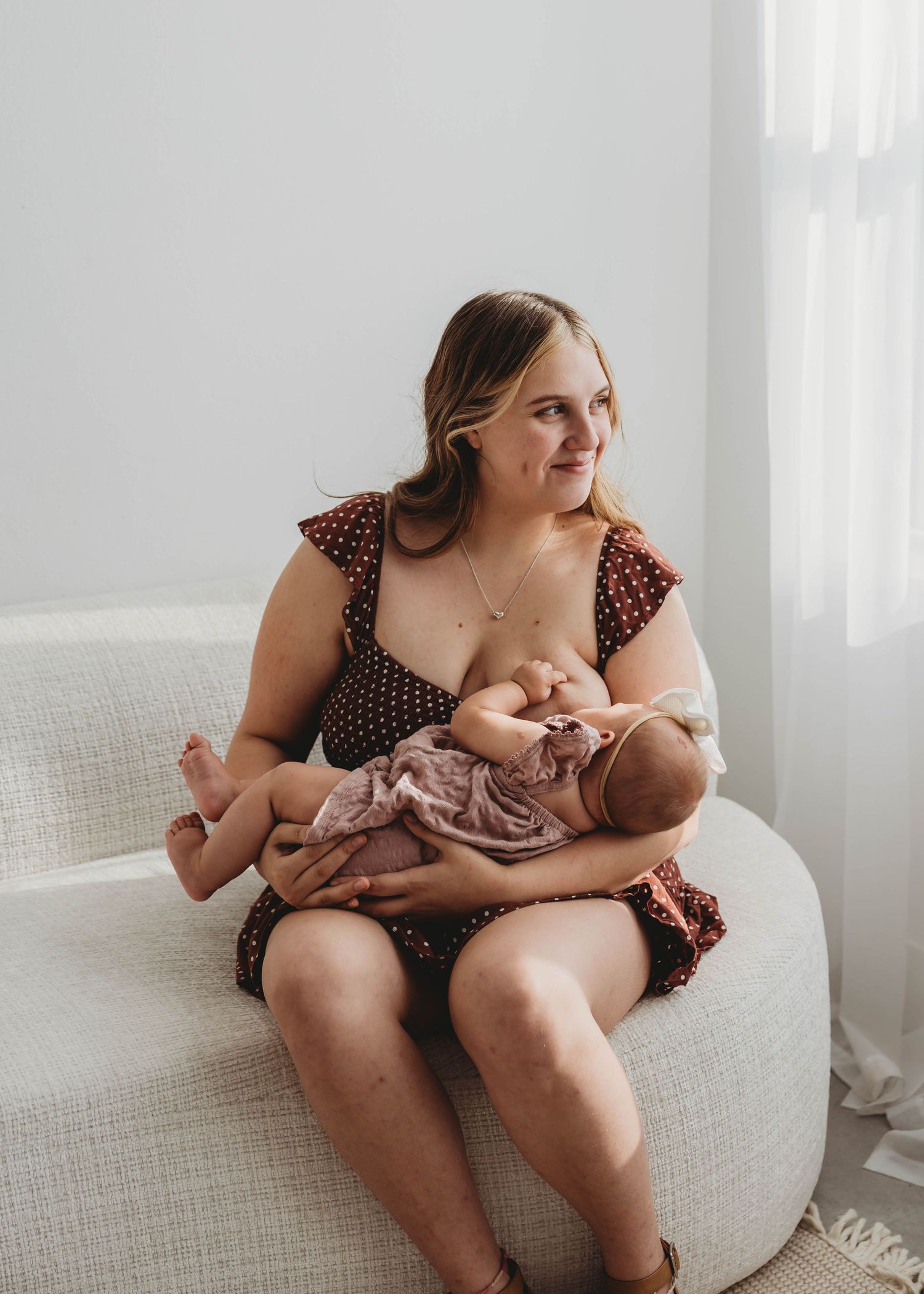 Mother in floral dress breastfeeding newborn baby while seated on white chair during in-home newborn session.