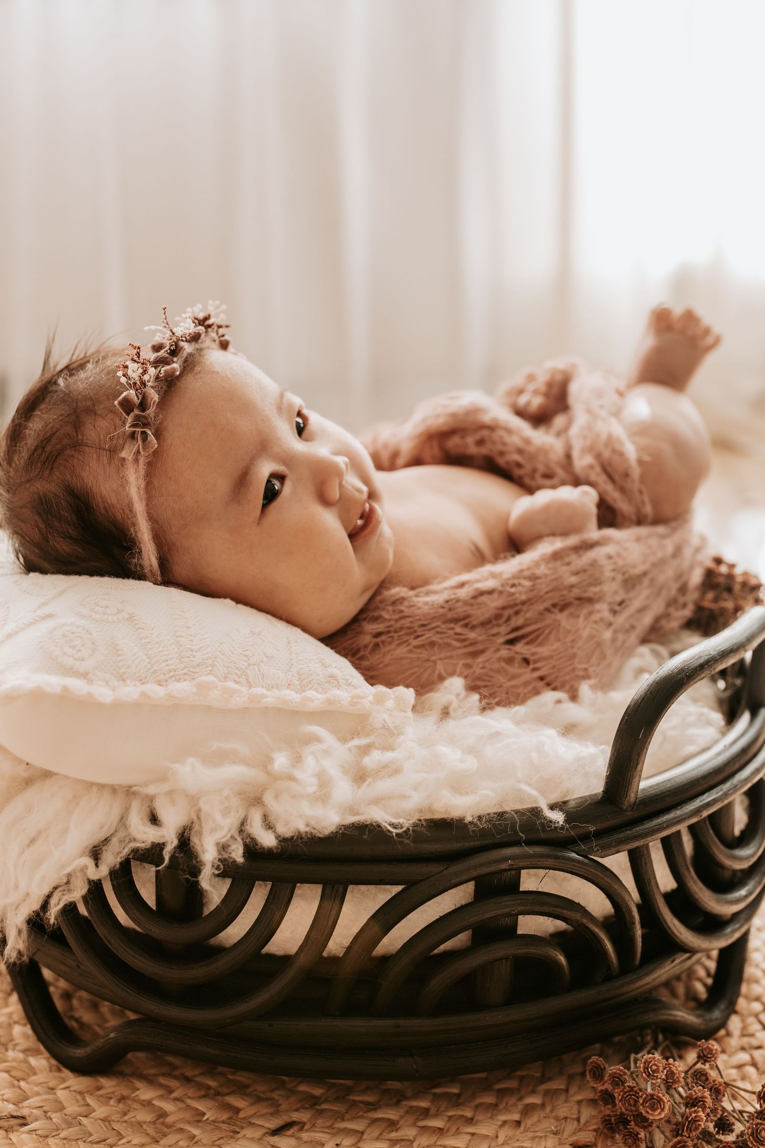 Newborn baby girl wearing a floral headband lying in a decorative rattan basket with soft white and pink blankets taken by Keely King
