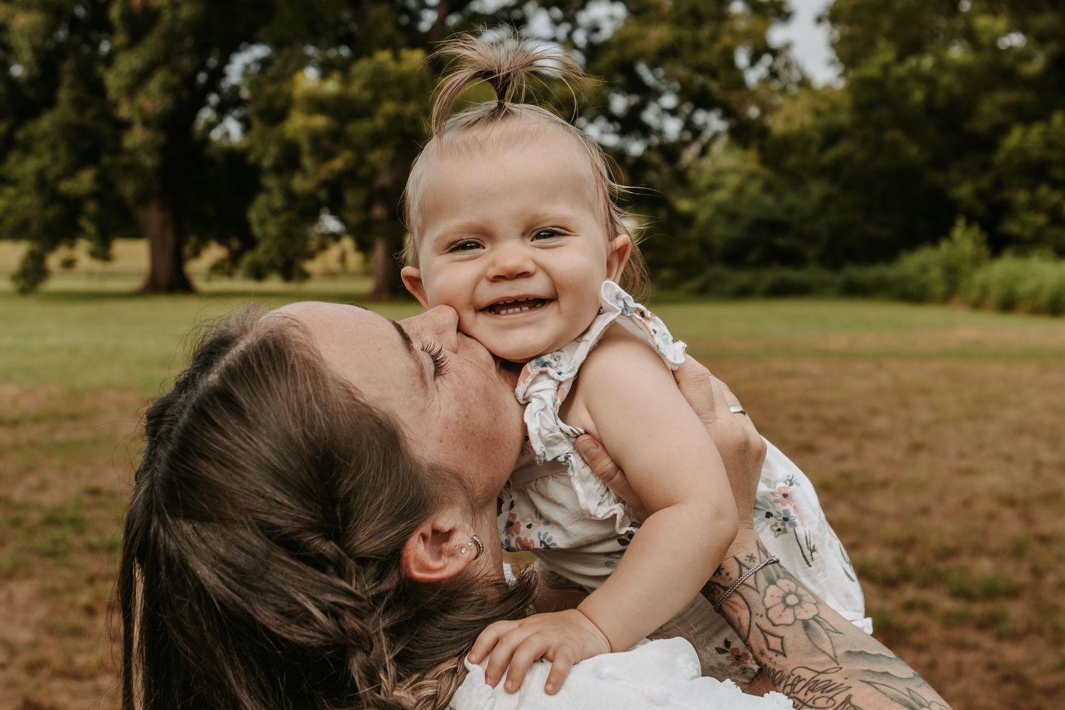 A mother lovingly kisses her smiling baby girl outdoors at Coal Creek Farm with Keely King Photography in Jenks Oklahoma