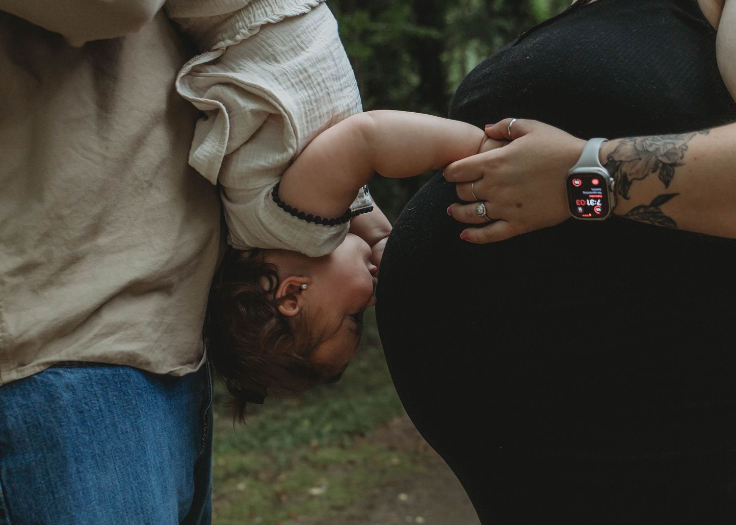 Pregnant woman and partner holding baby upside down outdoors, family maternity photo session in nature.