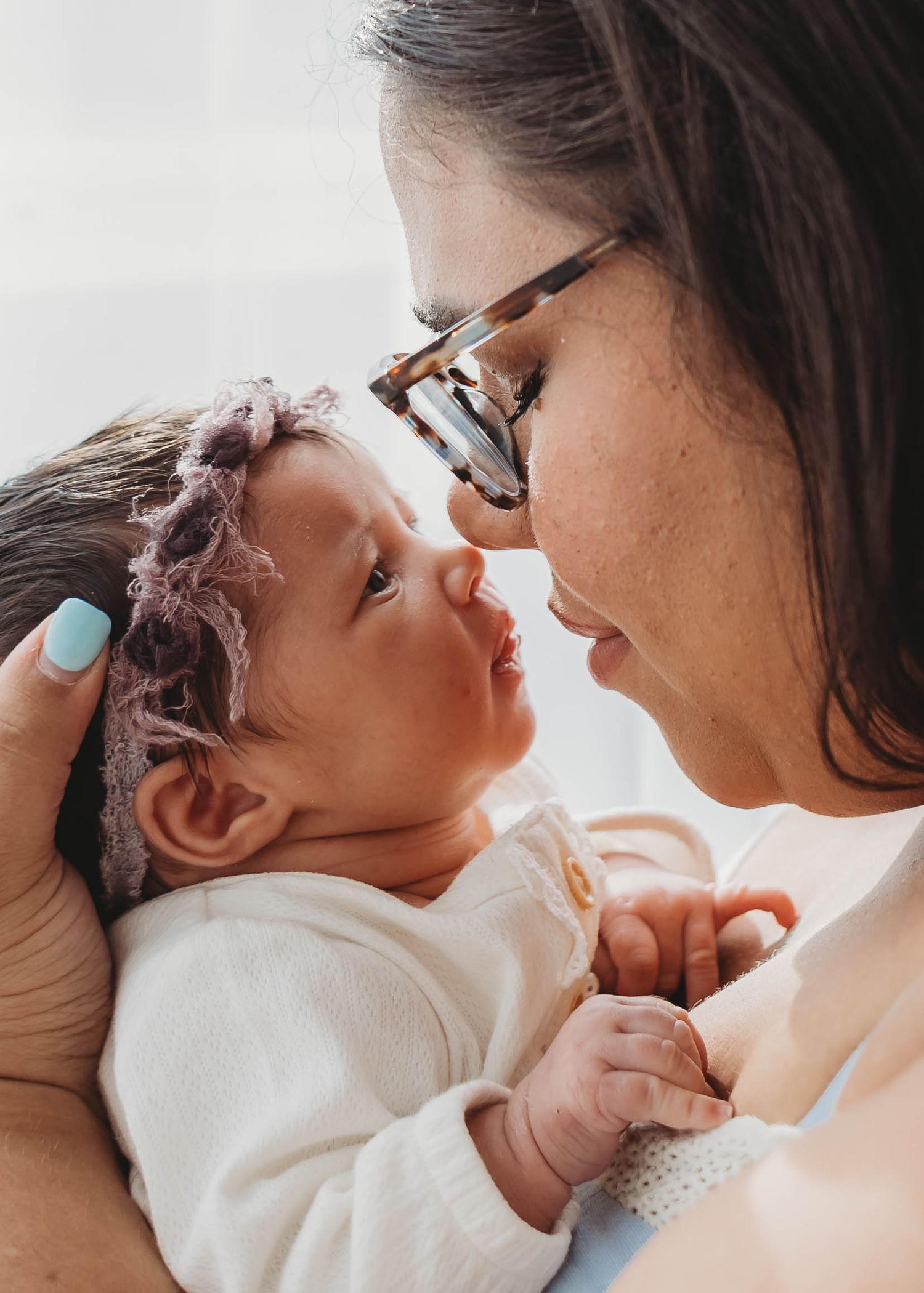 A mother with glasses tenderly holds her newborn baby, sharing a gentle nose-to-nose moment in soft natural light.
