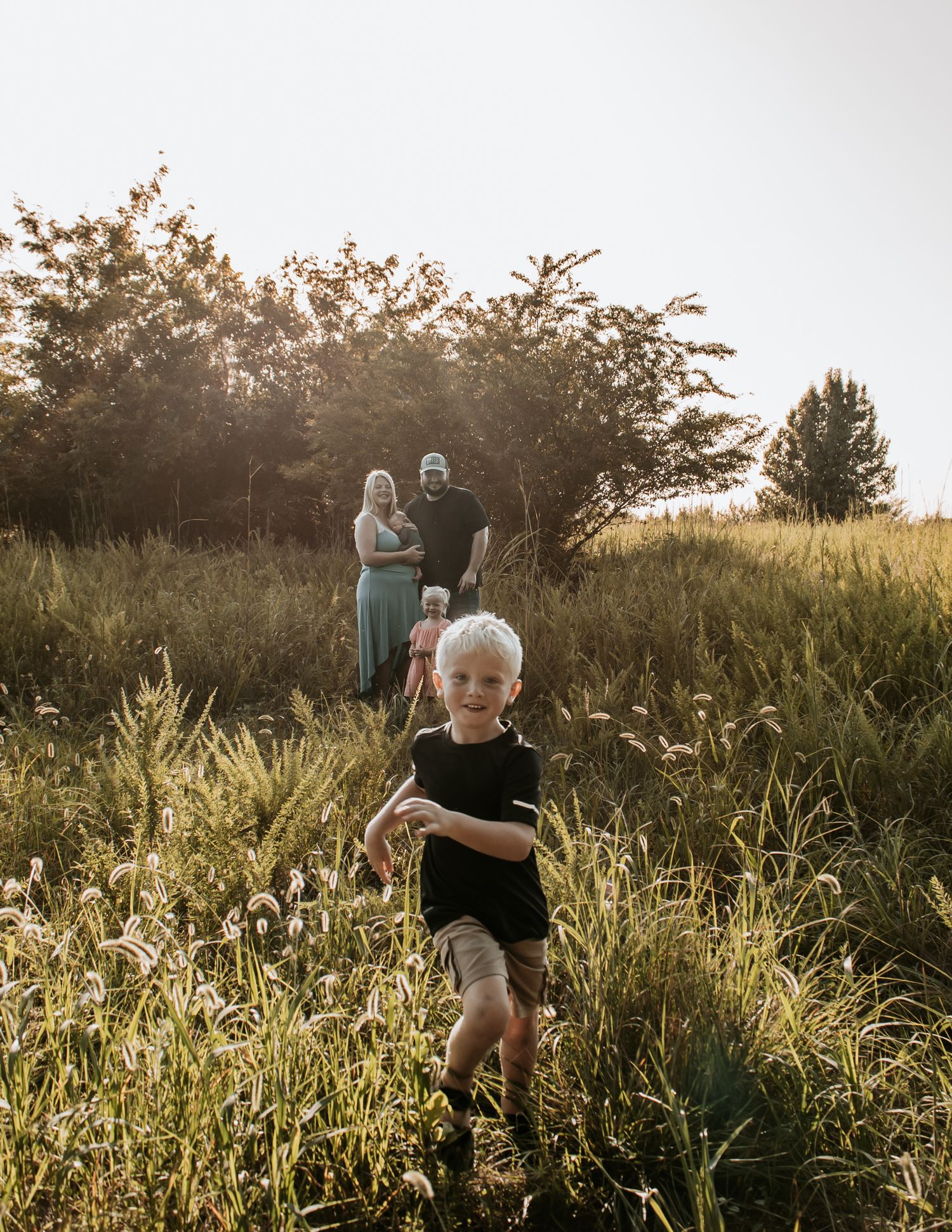 A young blonde boy runs through a sunny meadow while his family stands among tall grass and trees in the background in Claremore Oklahoma