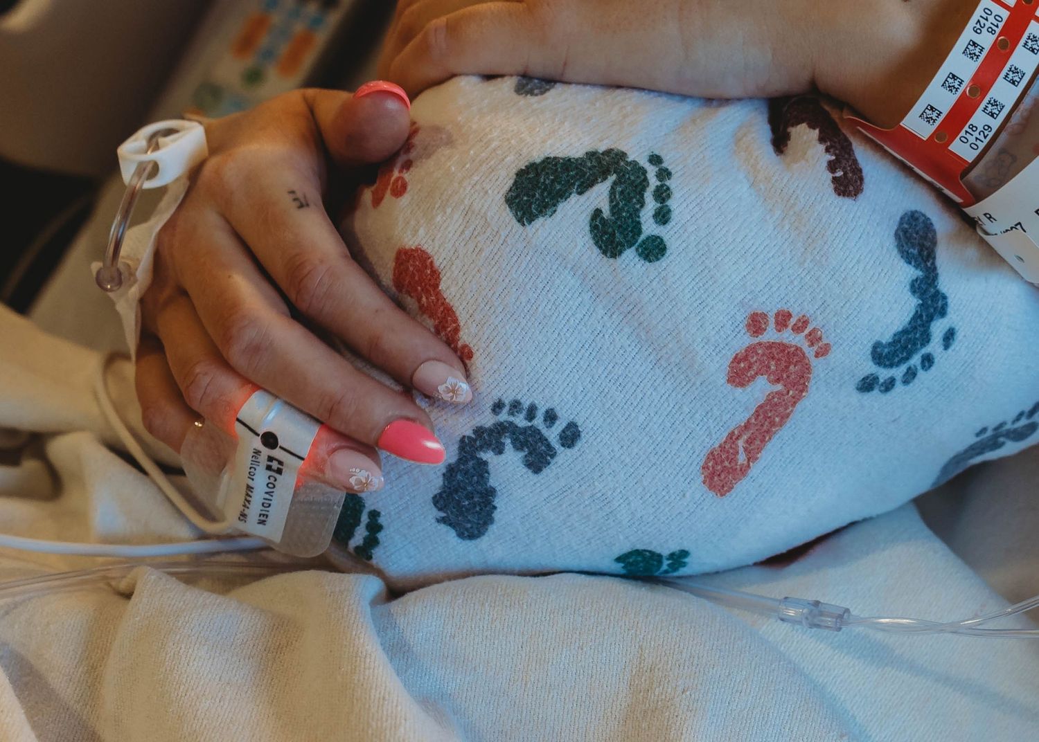 Mother with pink nails holds newborn baby wrapped in footprint blanket in hospital.