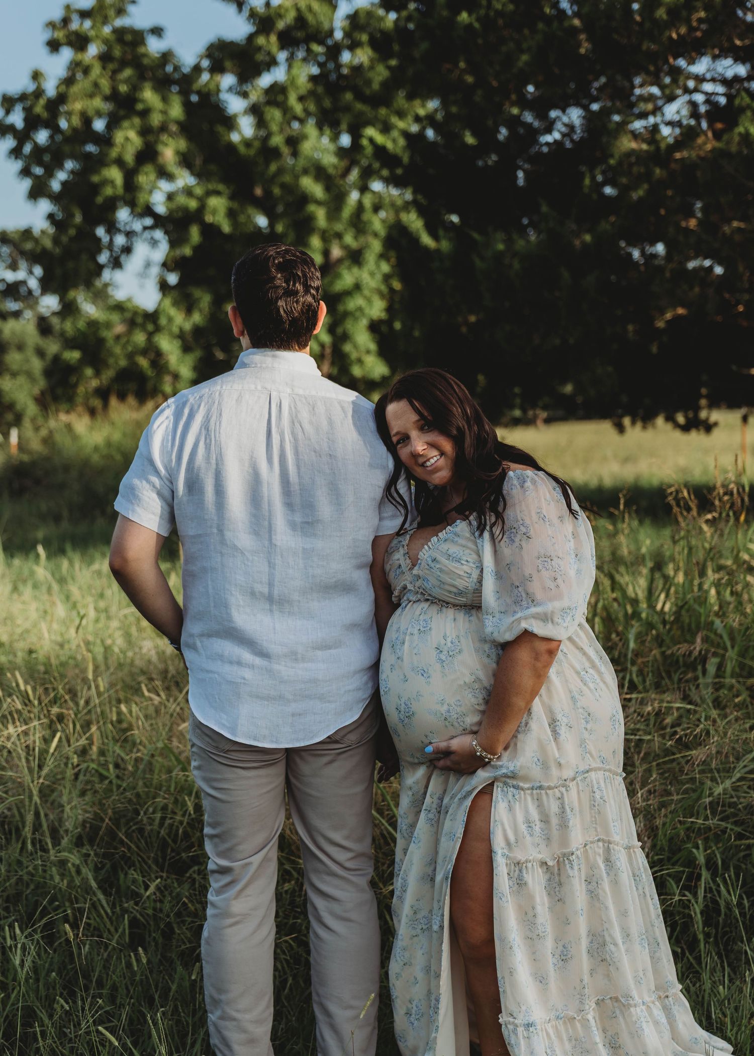 Pregnant woman in floral dress smiling at camera while standing with partner in green field.