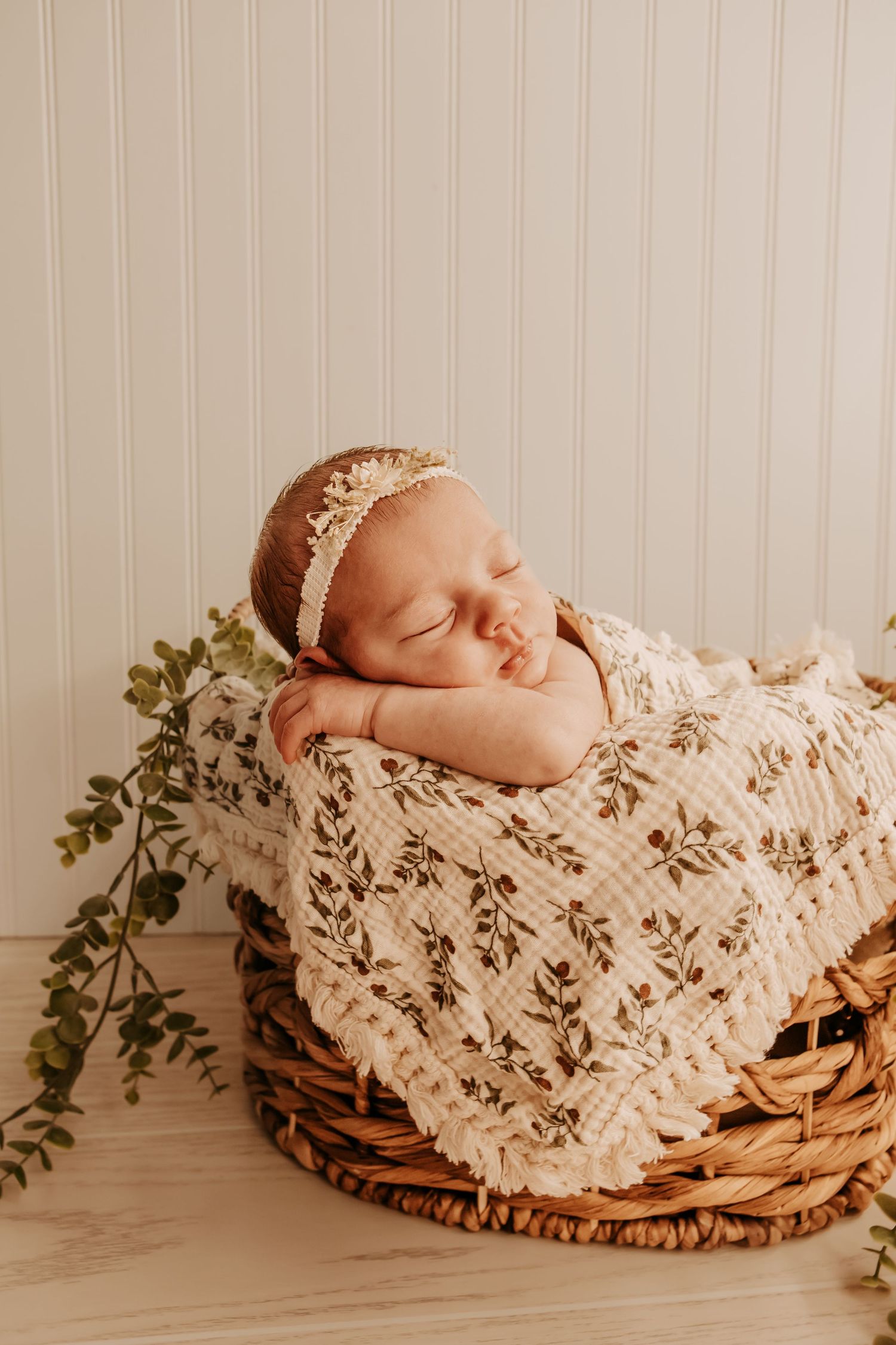 Sleeping newborn baby girl in floral wrap and lace headband nestled in wicker basket with greenery.