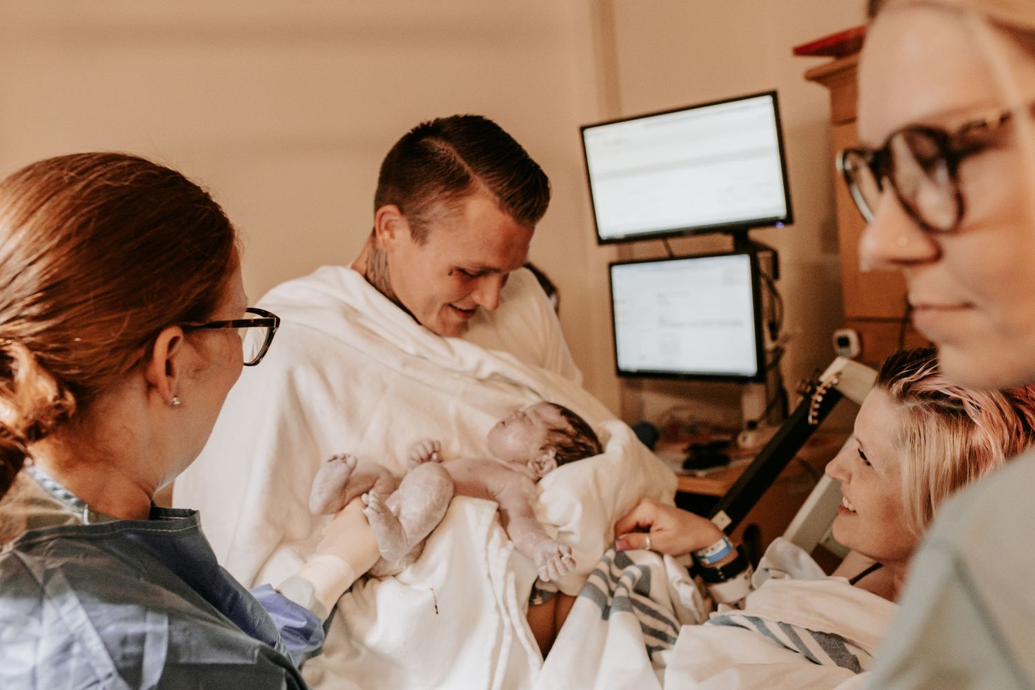 A father holds his newborn baby in a hospital room surrounded by medical staff and family after delivery.