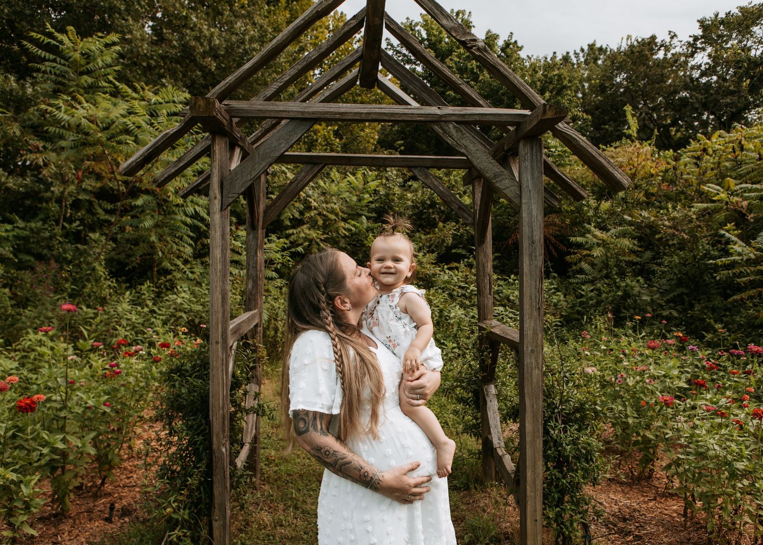 Pregnant mother kisses smiling child under a wooden arch surrounded by flowers with Keely King Photography in Jenks Oklahoma