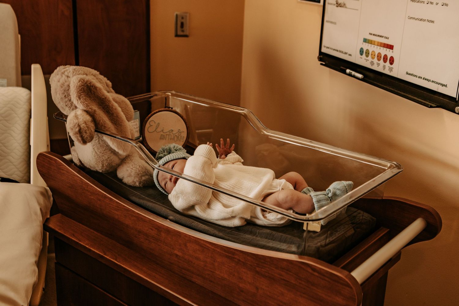 Newborn baby resting in hospital bassinet beside a stuffed bunny toy in a warm, softly lit delivery room.