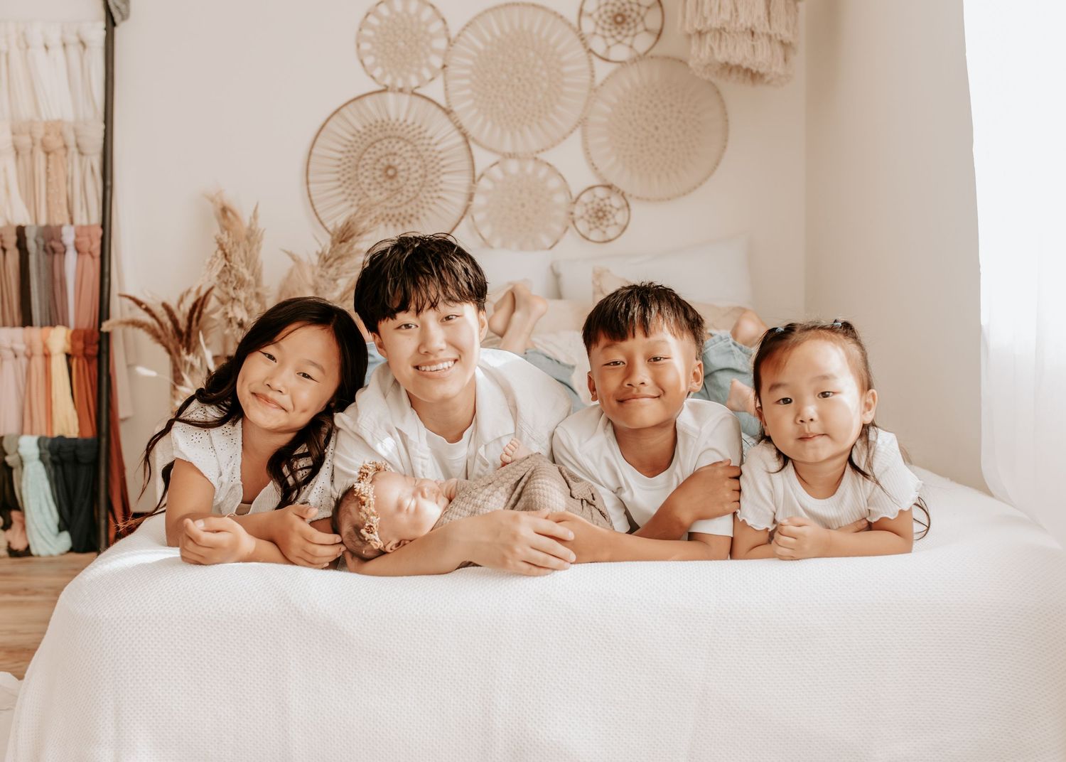 Four happy Asian children smiling and posing together on a white bed in a bright boho-style room.