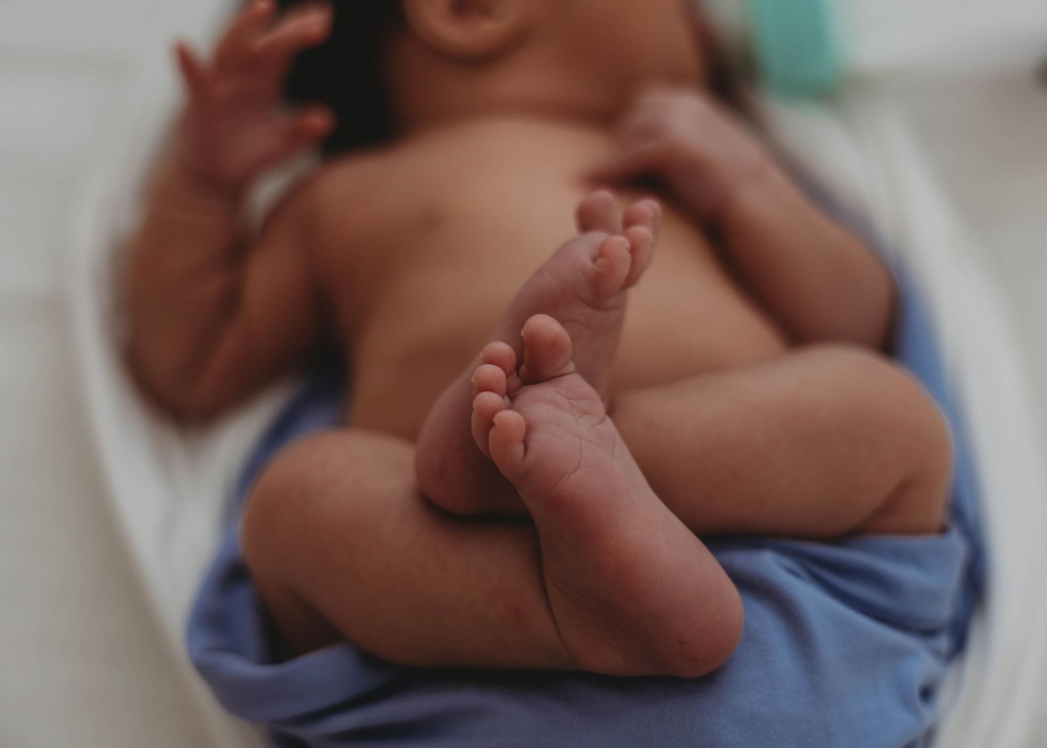 Newborn baby lying on a white surface wrapped in a blue blanket with tiny feet visible in the foreground.