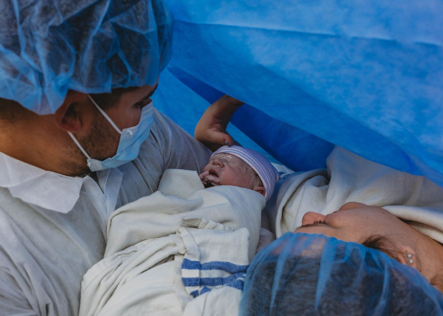 Father in surgical cap and mask cradles a newborn baby wrapped in a blanket in the operating room after a cesarean birth.