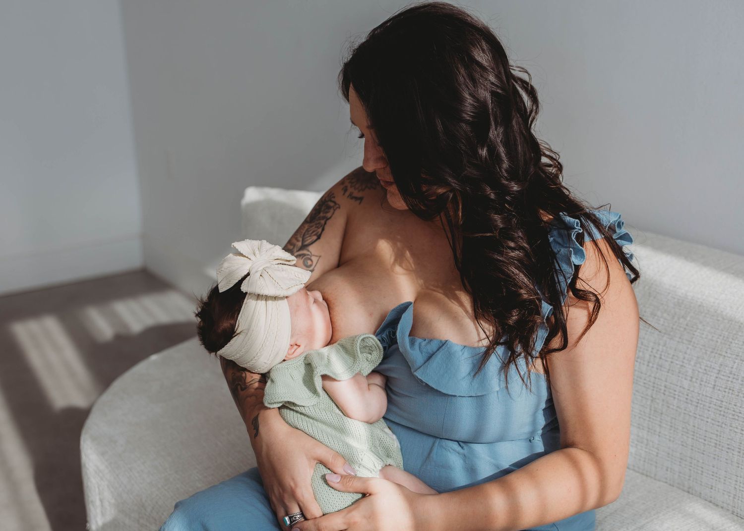 Dark-haired mother in blue dress nursing newborn with bow headband during a soft natural light newborn session.