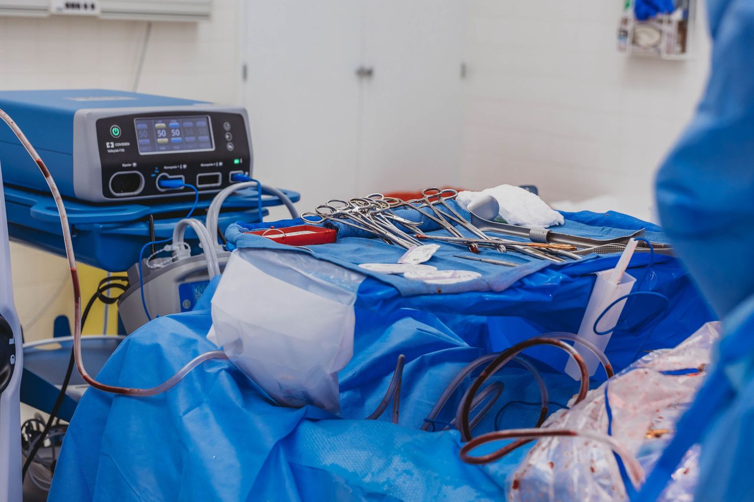 Surgical instruments and an electrosurgical unit rest on a blue sterile drape in an operating room, with a surgeon in blue scrubs nearby.