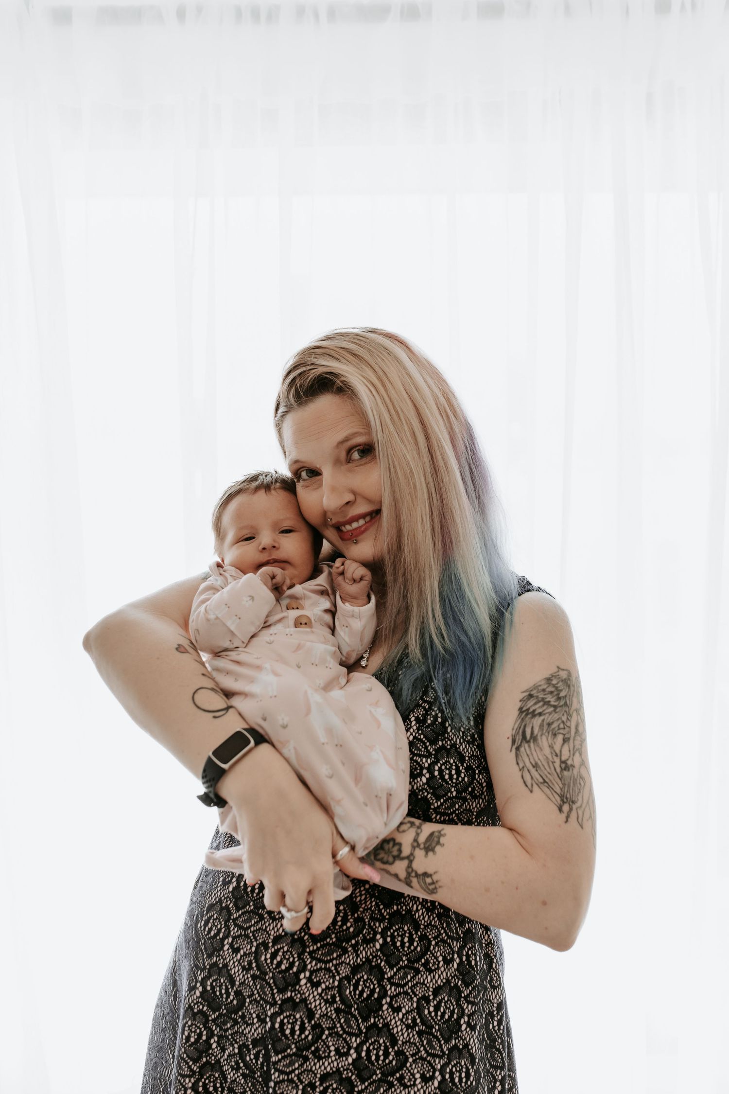 A smiling woman with blue-tipped hair and tattoos tenderly holds a newborn baby in a pink outfit against a white backdrop.