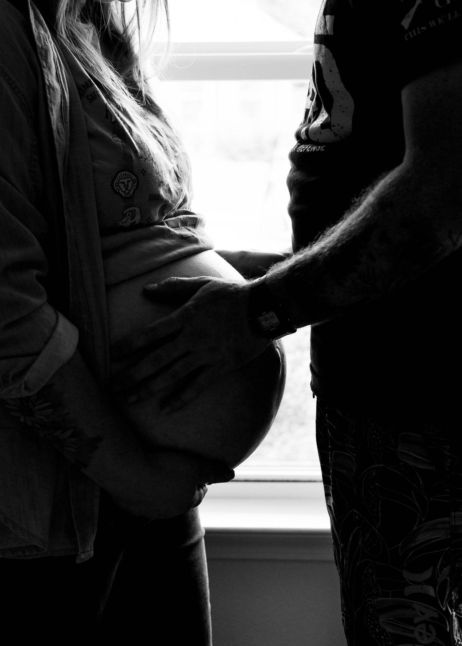 Black and white photo of a pregnant woman standing by a window with a partner gently touching her baby bump.