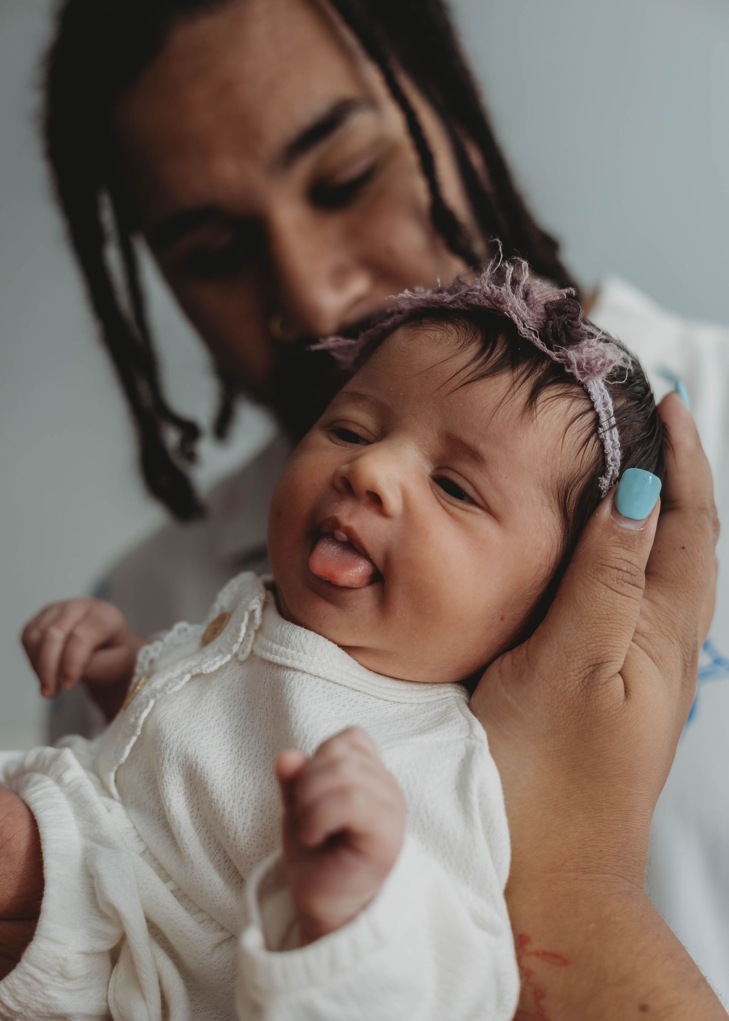 A father with dreadlocks tenderly holds a newborn baby girl wearing a pink headband and white onesie, sticking out her tongue.