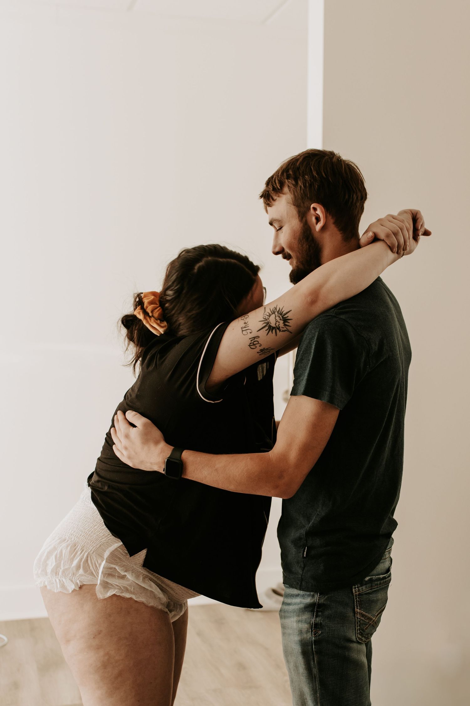 A couple embraces and smiles while dancing together, the man showing a sun tattoo on his arm.